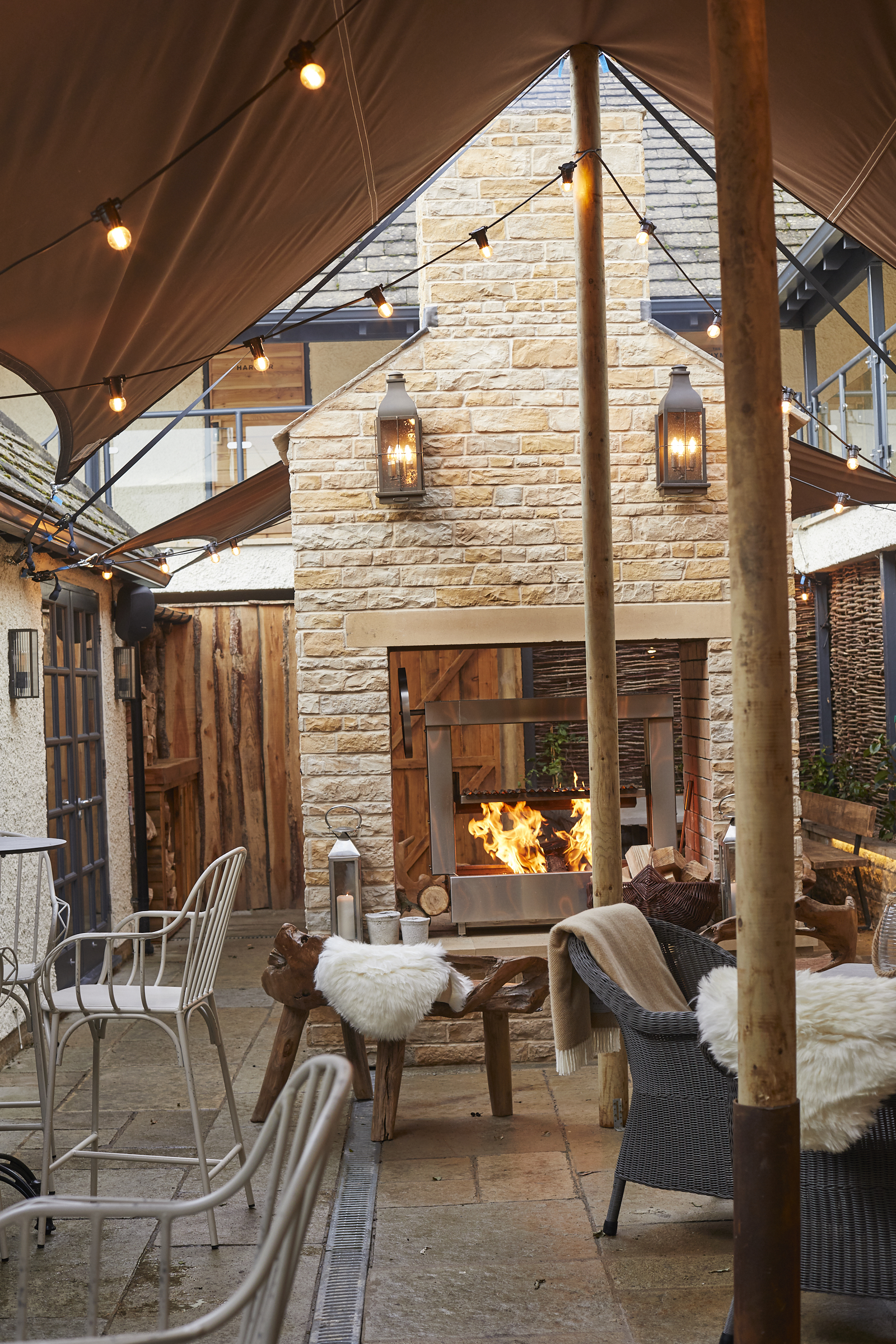 Cozy patio with a stone fireplace, decorated with string lights, wicker chairs, and wooden stools, under a tent canopy. - Wild Thyme & Honey Hotel in the Cotswolds