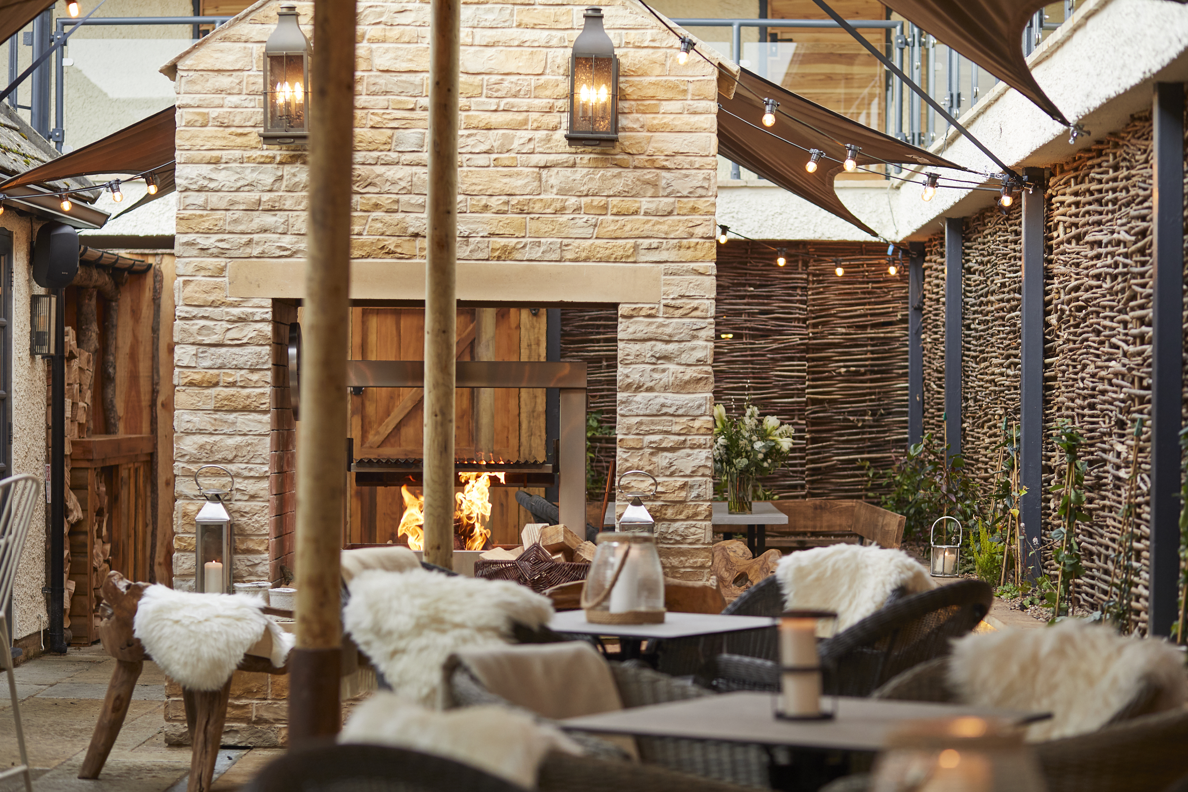 Cozy outdoor seating area with wicker chairs, soft furnishings, a stone fireplace, and ambient string lights. - Wild Thyme & Honey Hotel in the Cotswolds