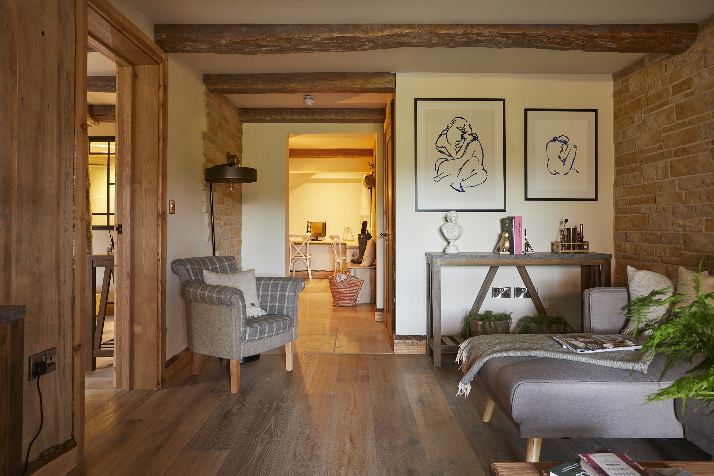 Cozy living room with wooden beams, gray furniture, framed art on walls. Hallway leads to a bright room with a table. - Wild Thyme & Honey Hotel in the Cotswolds