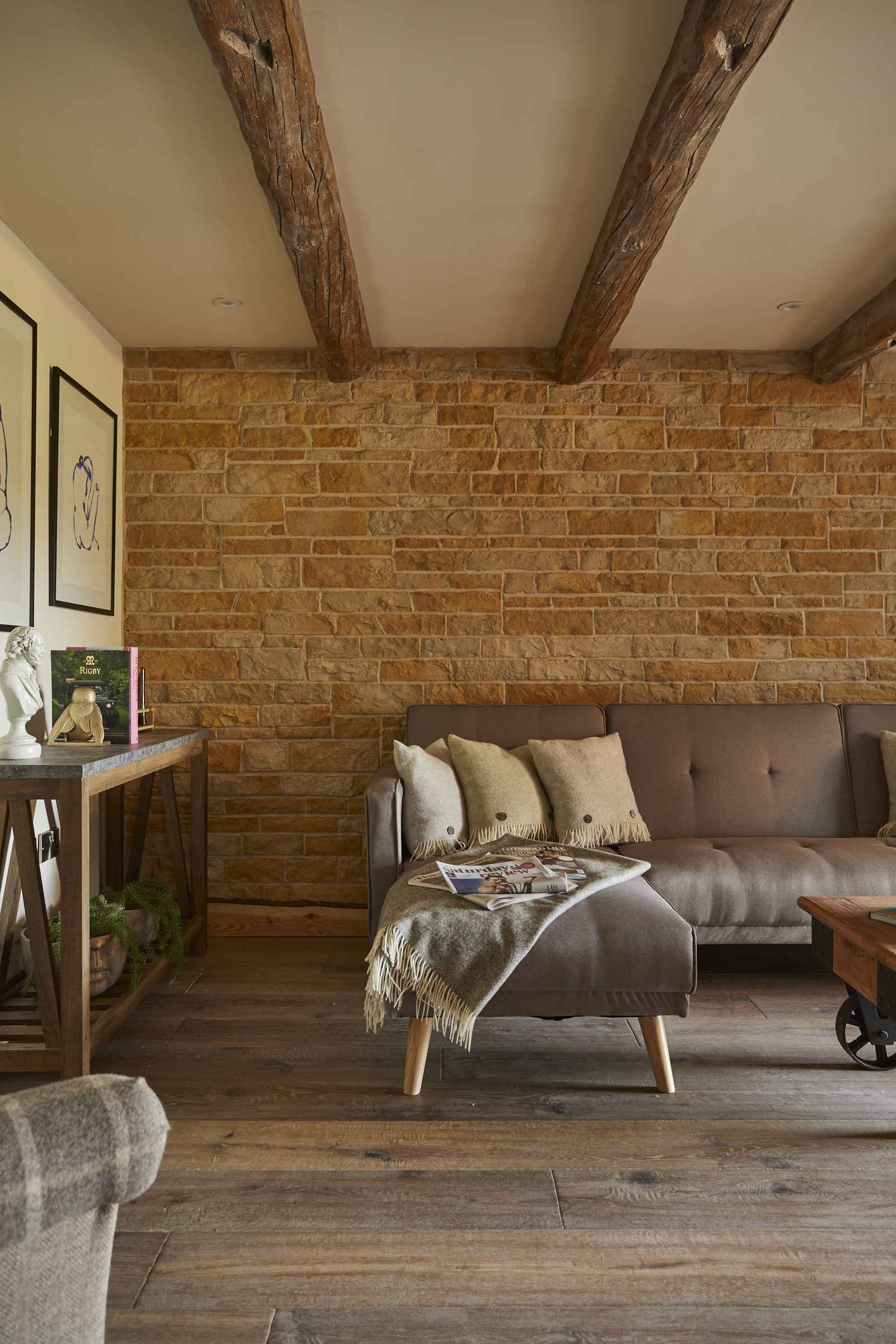 Cozy living room with wooden beams, stone wall, gray sofa, throw pillows, and a fringed blanket. Magazines on a coffee table. - Wild Thyme & Honey Hotel in the Cotswolds