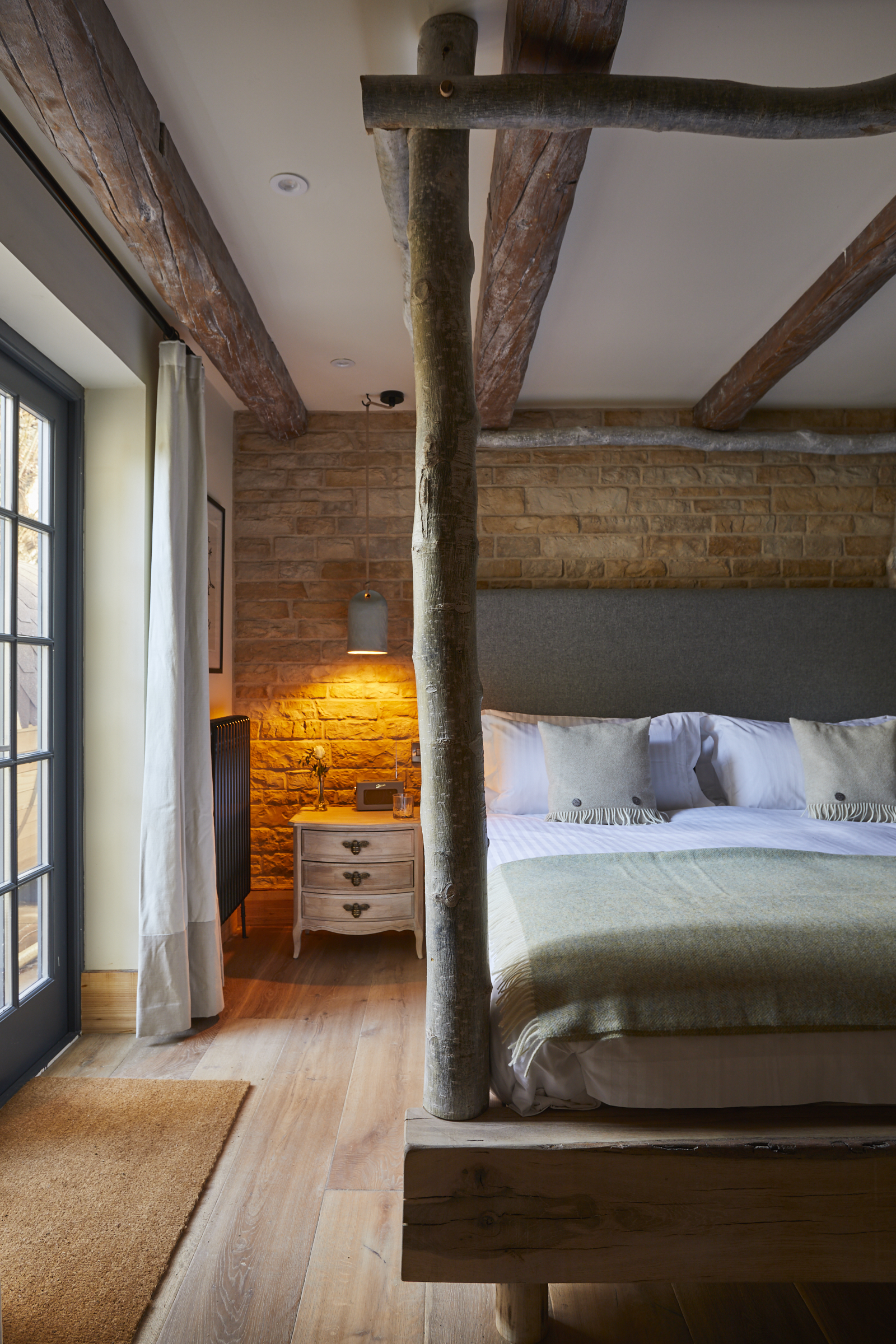 Bedroom with a rustic theme, featuring a wooden canopy bed, exposed wooden beams, and stone walls. A bedside table with a lit lamp stands beside the bed, and large windows with curtains allow natural light into the room. Warm, cozy atmosphere. - Wild Thyme & Honey Hotel in the Cotswolds