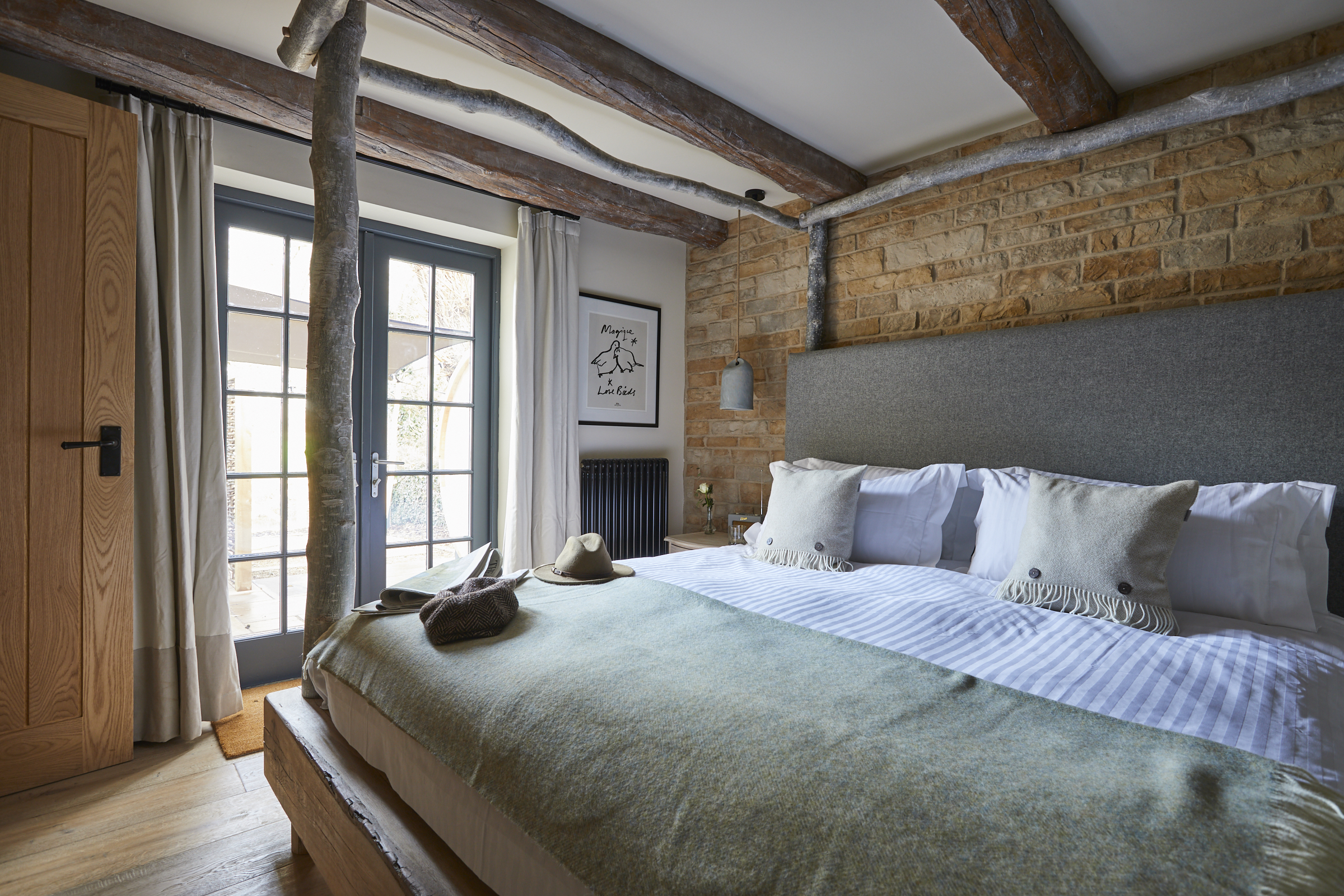 A cozy bedroom with a large bed, plaid comforter, wooden beams, exposed brick wall, and glass door leading to an outside area. - Wild Thyme & Honey Hotel in the Cotswolds