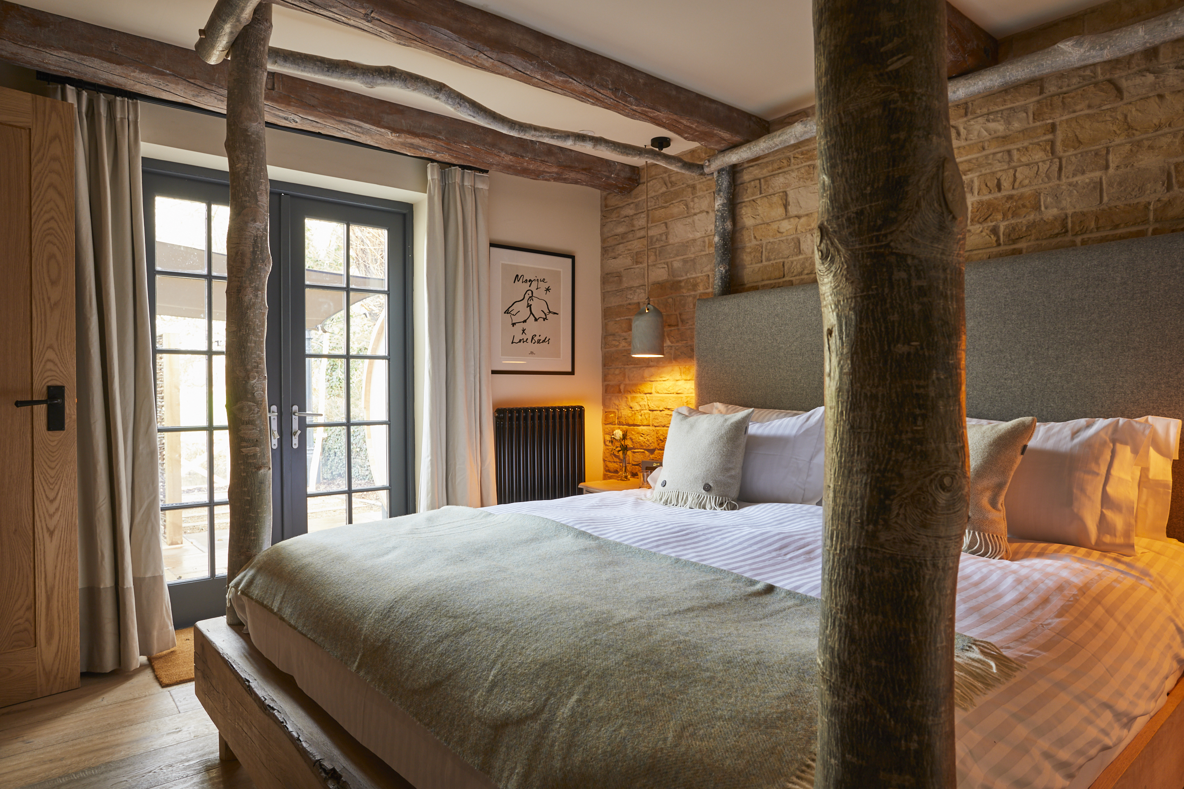 Cozy bedroom with a rustic, log bed frame, a large window, brick walls, and soft lighting. Art hangs above the bed. - Wild Thyme & Honey Hotel in the Cotswolds