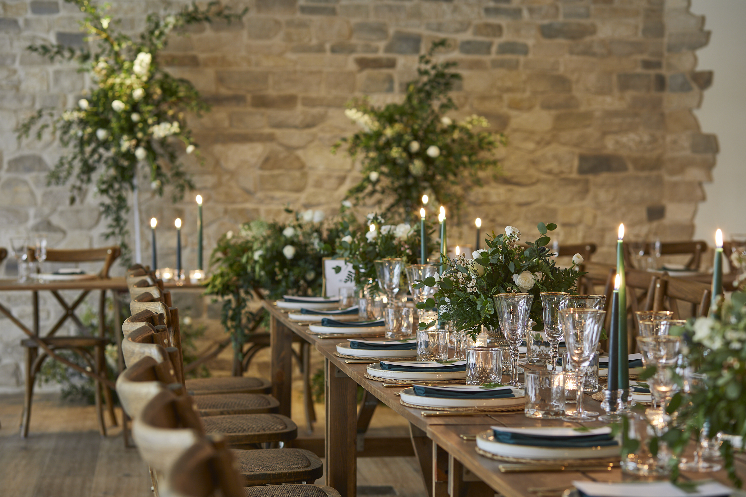 Elegant banquet table set with green and white decor, candles, glassware, and floral arrangements against a stone wall backdrop. - Wild Thyme & Honey Hotel in the Cotswolds