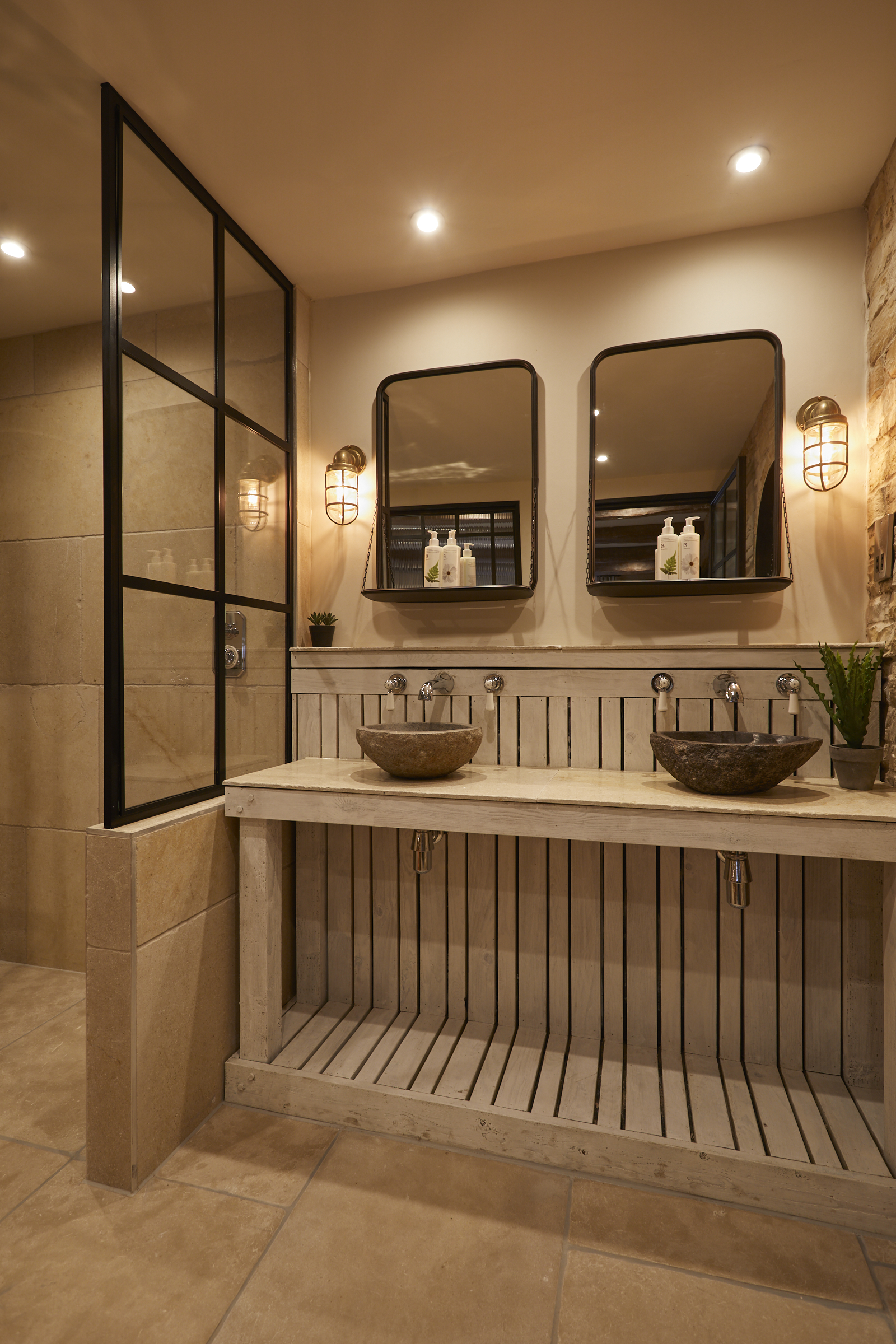 Modern bathroom with two stone sinks, rectangular mirrors, and wall sconces. Glass shower door and wooden counter. Warm lighting. - Wild Thyme & Honey Hotel in the Cotswolds