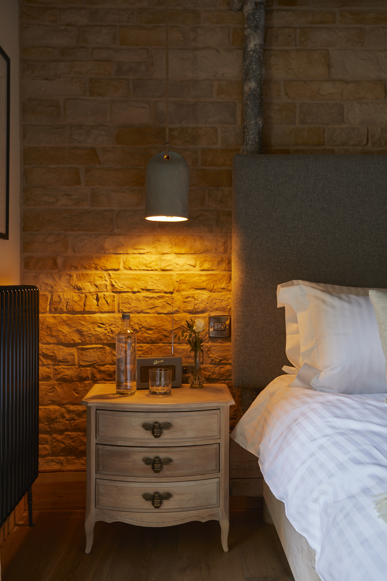 Cozy bedroom with a wooden bedside table, lit pendant light, brick wall, and neatly made bed with white linens. - Wild Thyme & Honey Hotel in the Cotswolds
