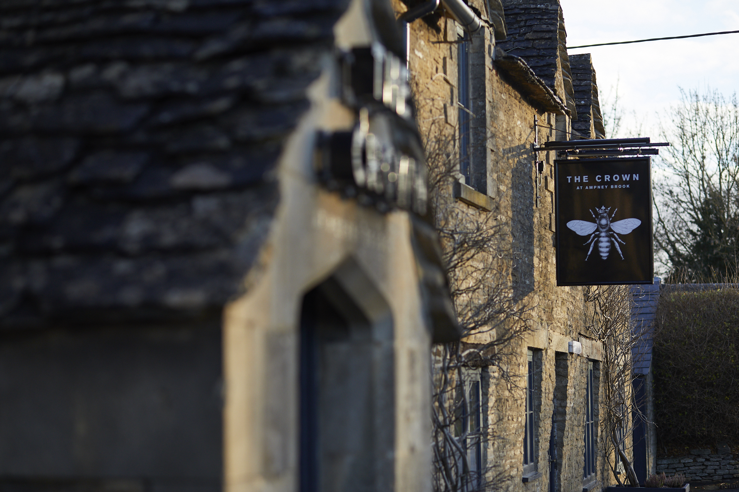 Stone buildings with a pub sign reading The Crown featuring a bee; the scene is lit by soft natural light. - Wild Thyme & Honey Hotel in the Cotswolds