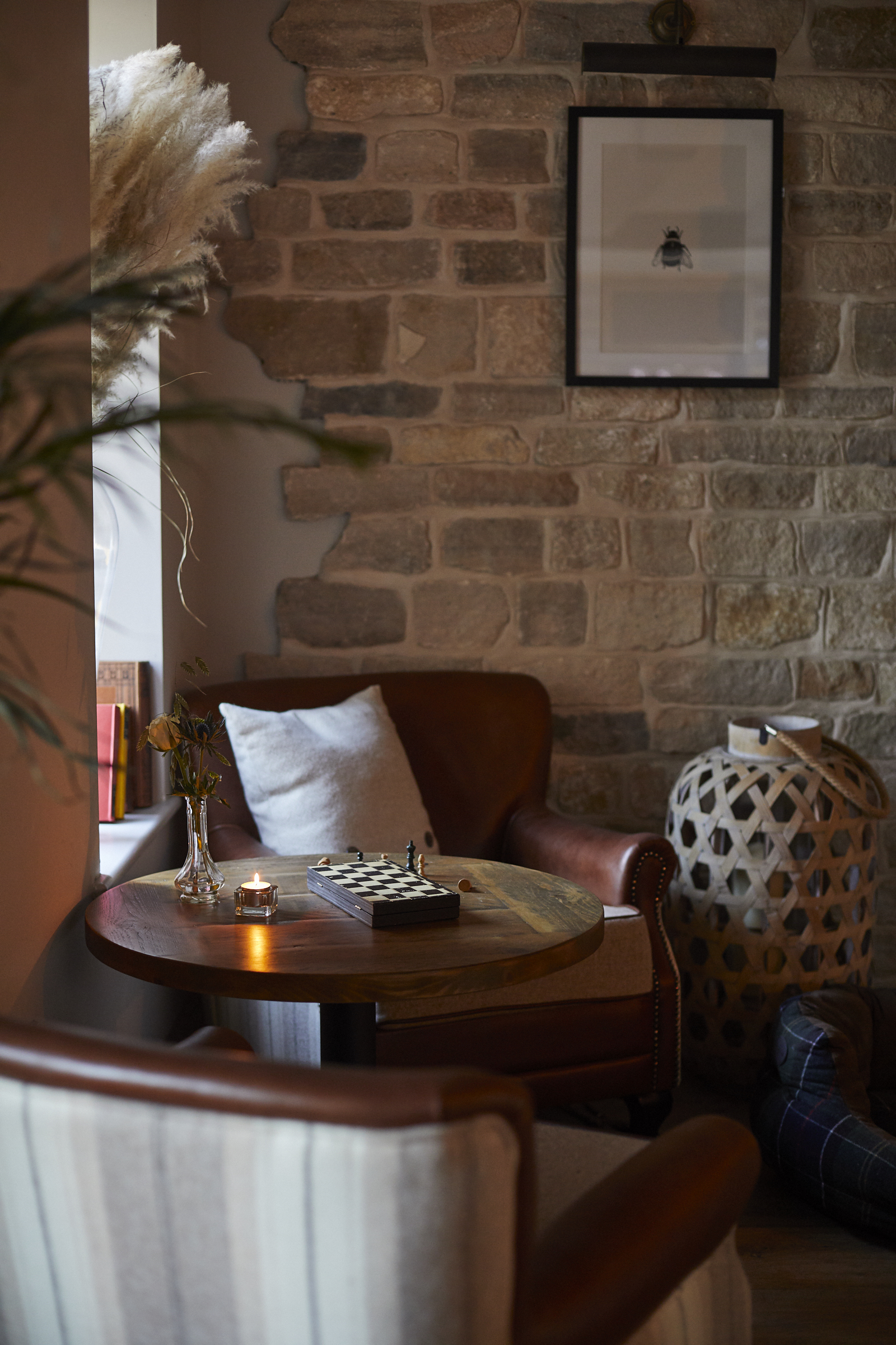 Cozy nook with leather chairs, round wooden table, chessboard, candle, framed picture, and stone wall in soft lighting. - Wild Thyme & Honey Hotel in the Cotswolds