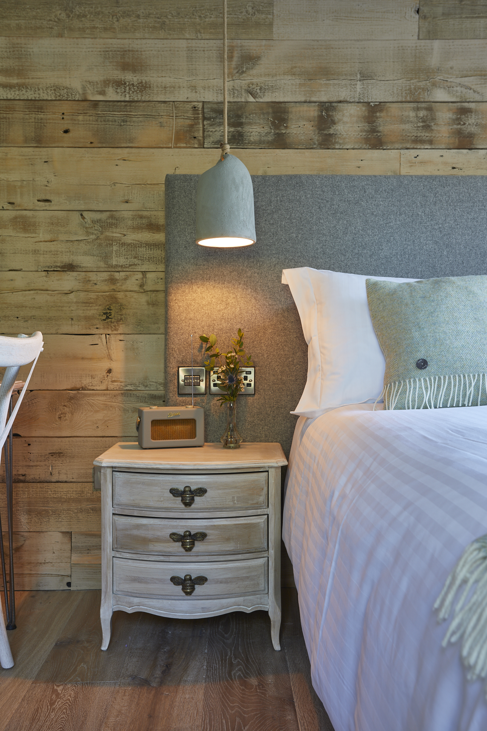 Cozy bedroom with a wooden nightstand, a pendant light, vintage radio, and a neatly made bed with gray and white bedding. - Wild Thyme & Honey Hotel in the Cotswolds