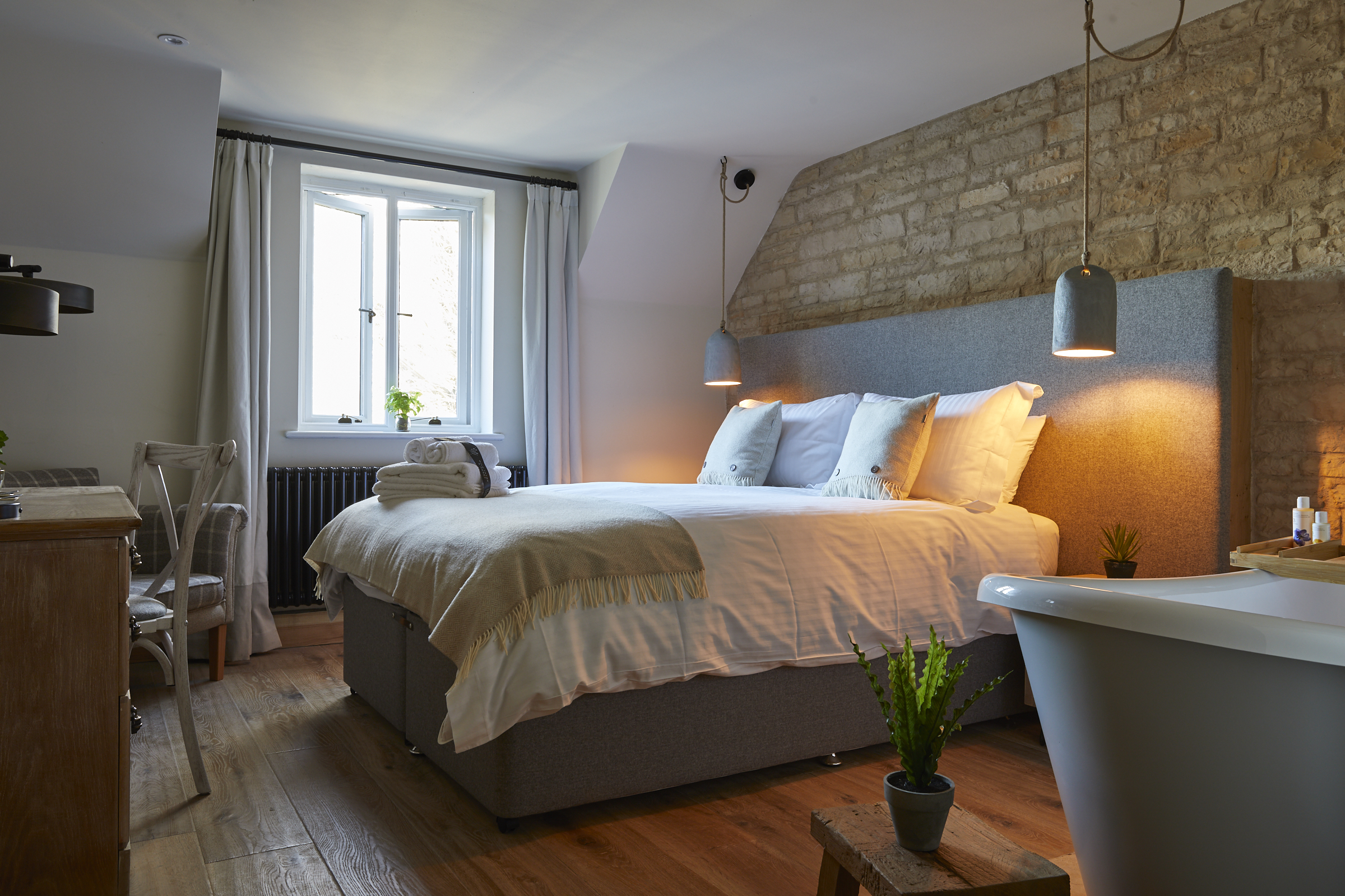 Cozy bedroom with a double bed, pillows, bedside lamps, a window, wooden floor, chairs, and a bathtub in the foreground. - Wild Thyme & Honey Hotel in the Cotswolds