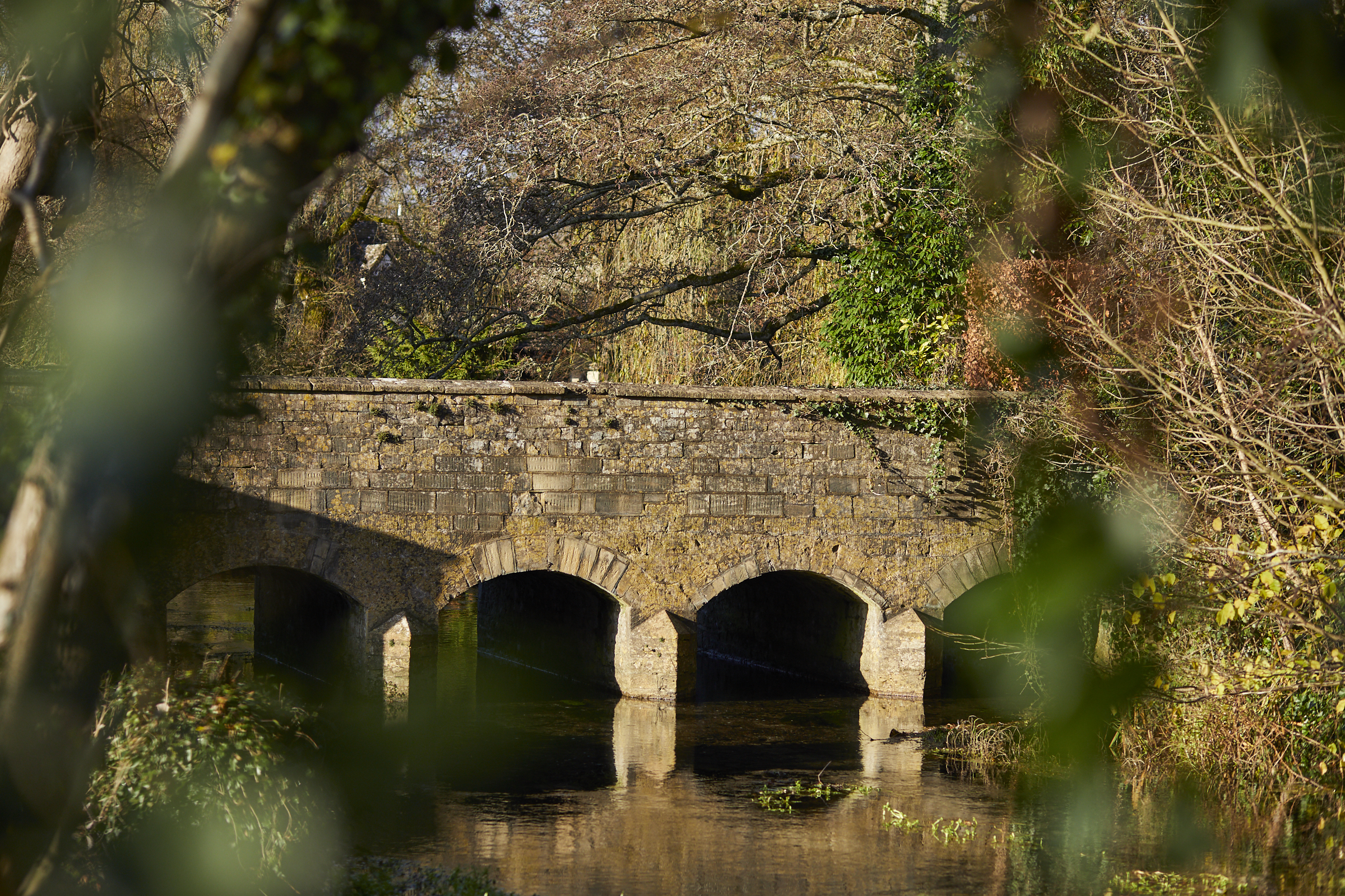 Stone bridge with three arches over a calm river, surrounded by lush greenery and trees in a serene, natural setting. - Wild Thyme & Honey Hotel in the Cotswolds