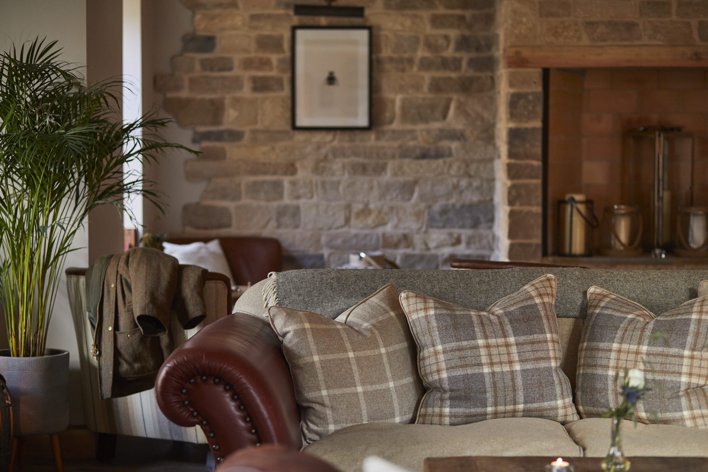 Cozy living room with a plaid-cushioned sofa, hanging jacket, potted plant, and stone wall in the background. - Wild Thyme & Honey Hotel in the Cotswolds