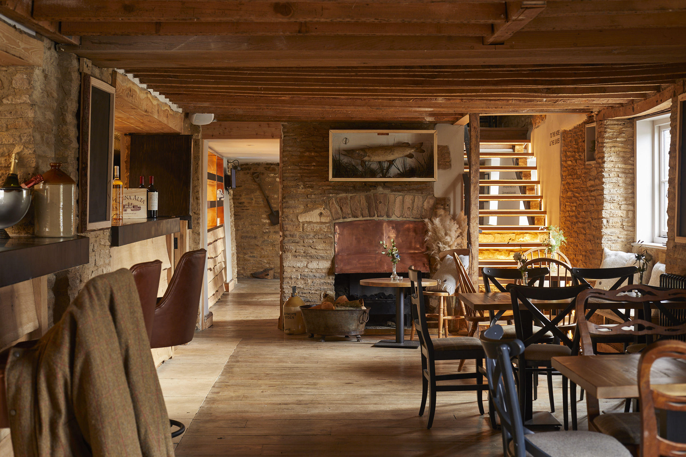 Cozy rustic café interior with wooden beams and stone walls. Tables and chairs are arranged around the room. A fireplace with seating, shelves with bottles, and a staircase are visible. Soft natural light comes in through a window. - Wild Thyme & Honey Hotel in the Cotswolds