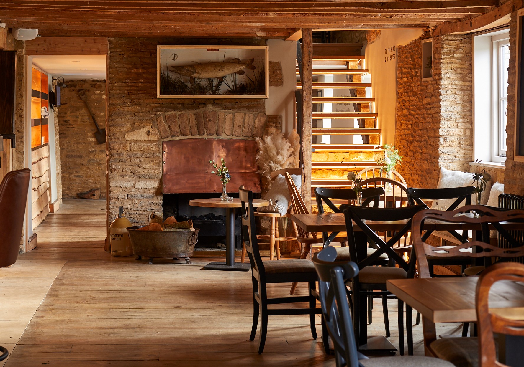 Cozy rustic cafe with wooden tables, chairs, exposed beams, stone walls, and a fireplace, lit by natural light from a window. - Wild Thyme & Honey Hotel in the Cotswolds