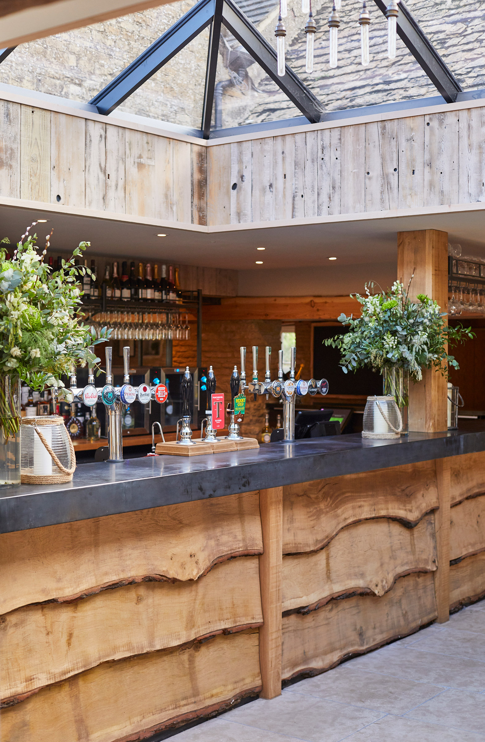 Rustic bar interior with a wooden counter and beer taps. Greenery in vases decorates the counter. A skylight above lets in natural light, highlighting the stone walls and wooden elements. Shelves with bottles are visible in the background. - Wild Thyme & Honey Hotel in the Cotswolds