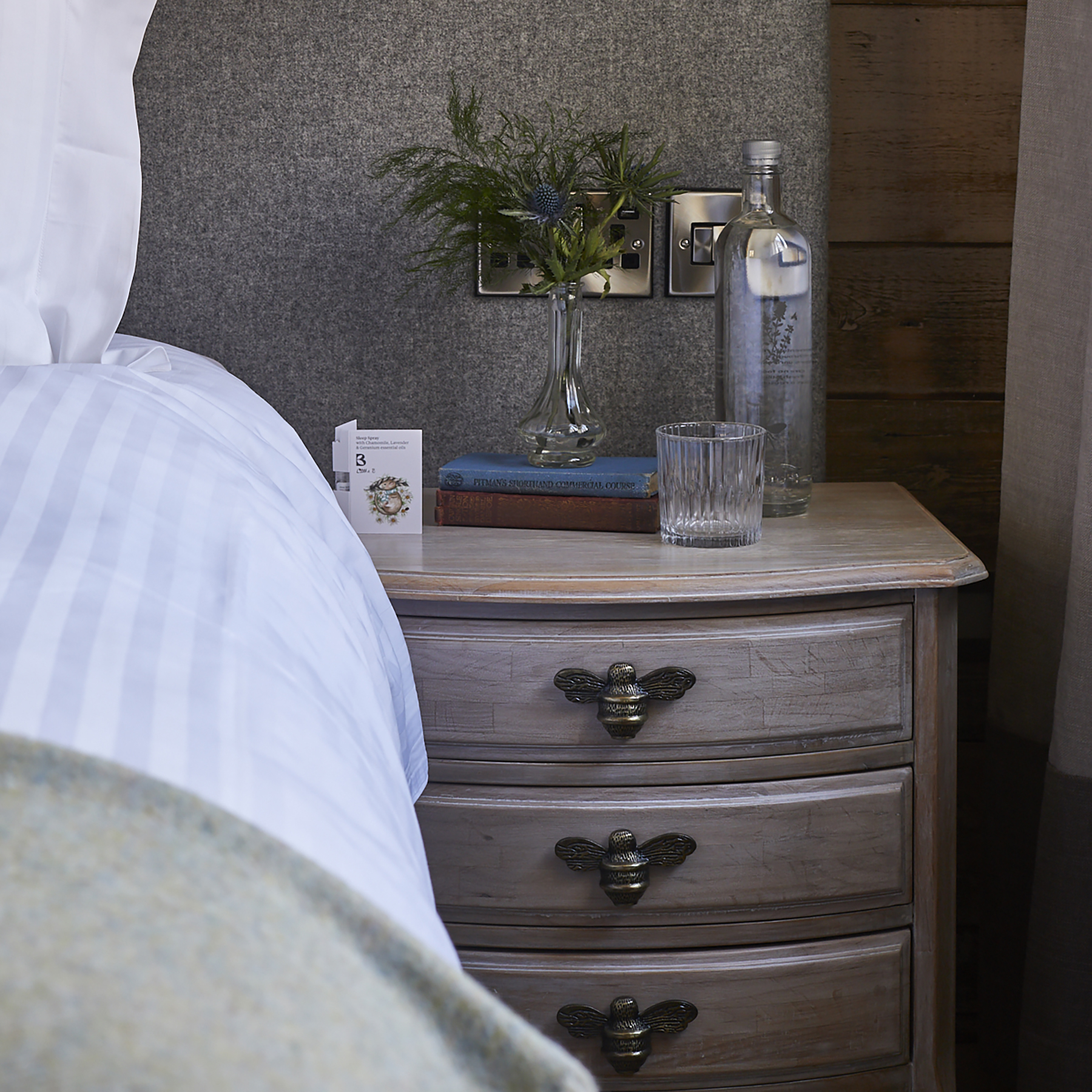 A cozy bedroom nightstand with a water bottle, glass, books, a small flower vase, and a box of tissues beside a bed. - Wild Thyme & Honey Hotel in the Cotswolds