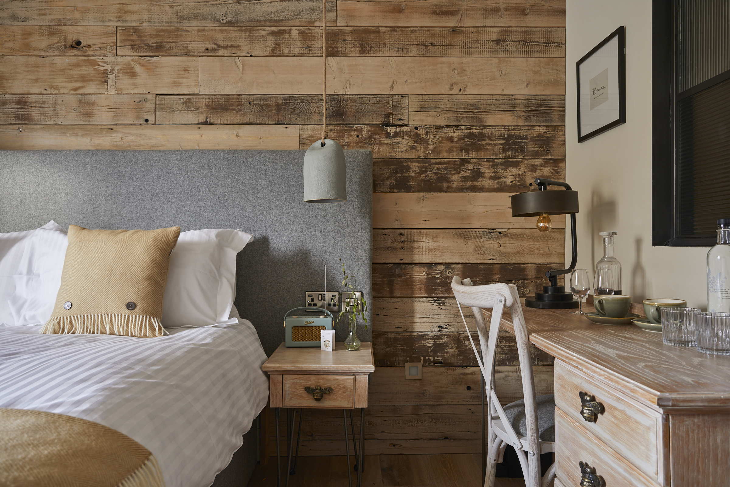 Cozy bedroom with rustic wood accent wall, gray upholstered headboard, desk, chair, pendant lamp, and decorative bedside items. - Wild Thyme & Honey Hotel in the Cotswolds