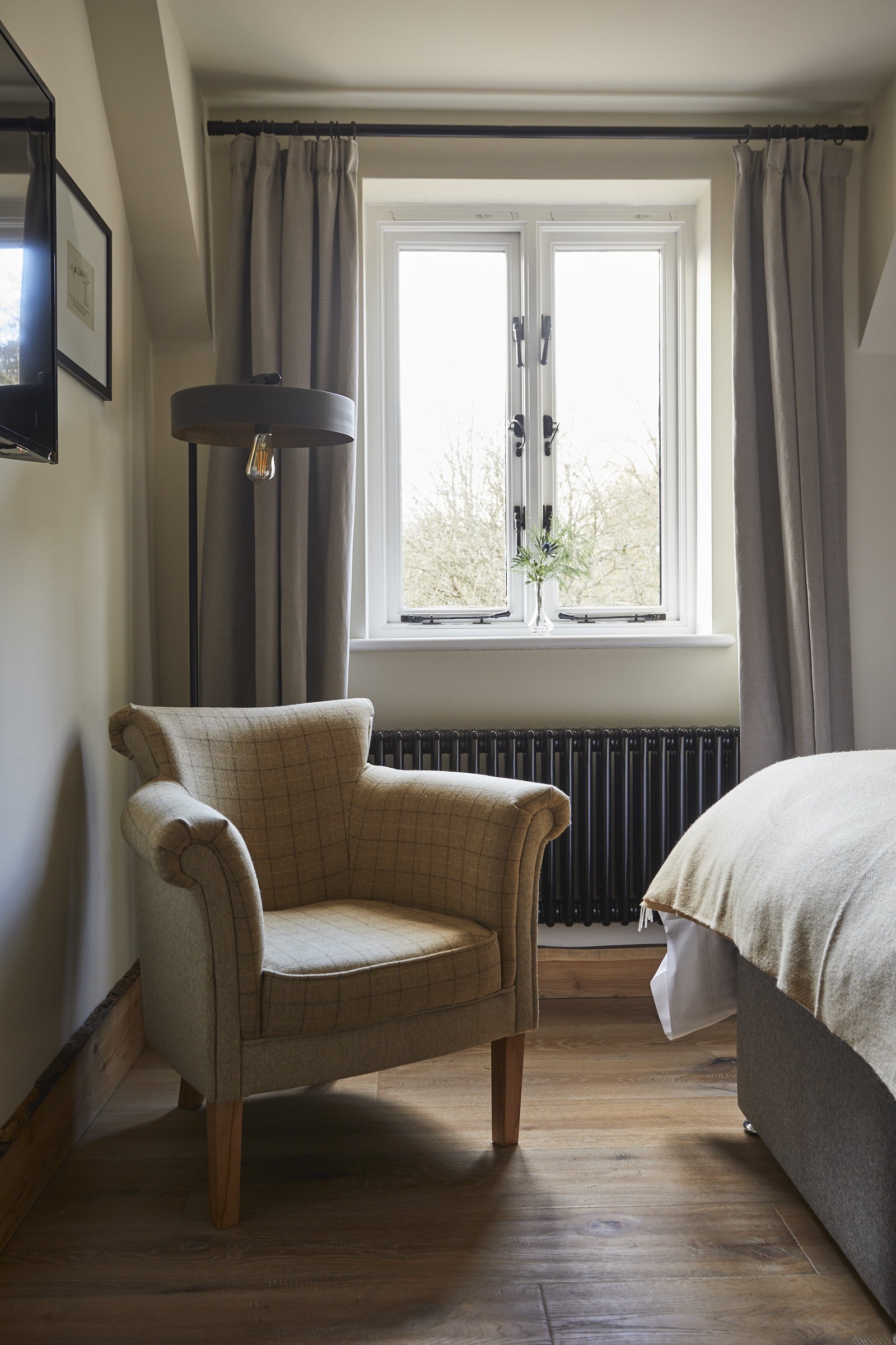 Cozy room with an armchair by the window, curtains drawn, and a lamp overhead. Bed visible with light-colored linens. - Wild Thyme & Honey Hotel in the Cotswolds
