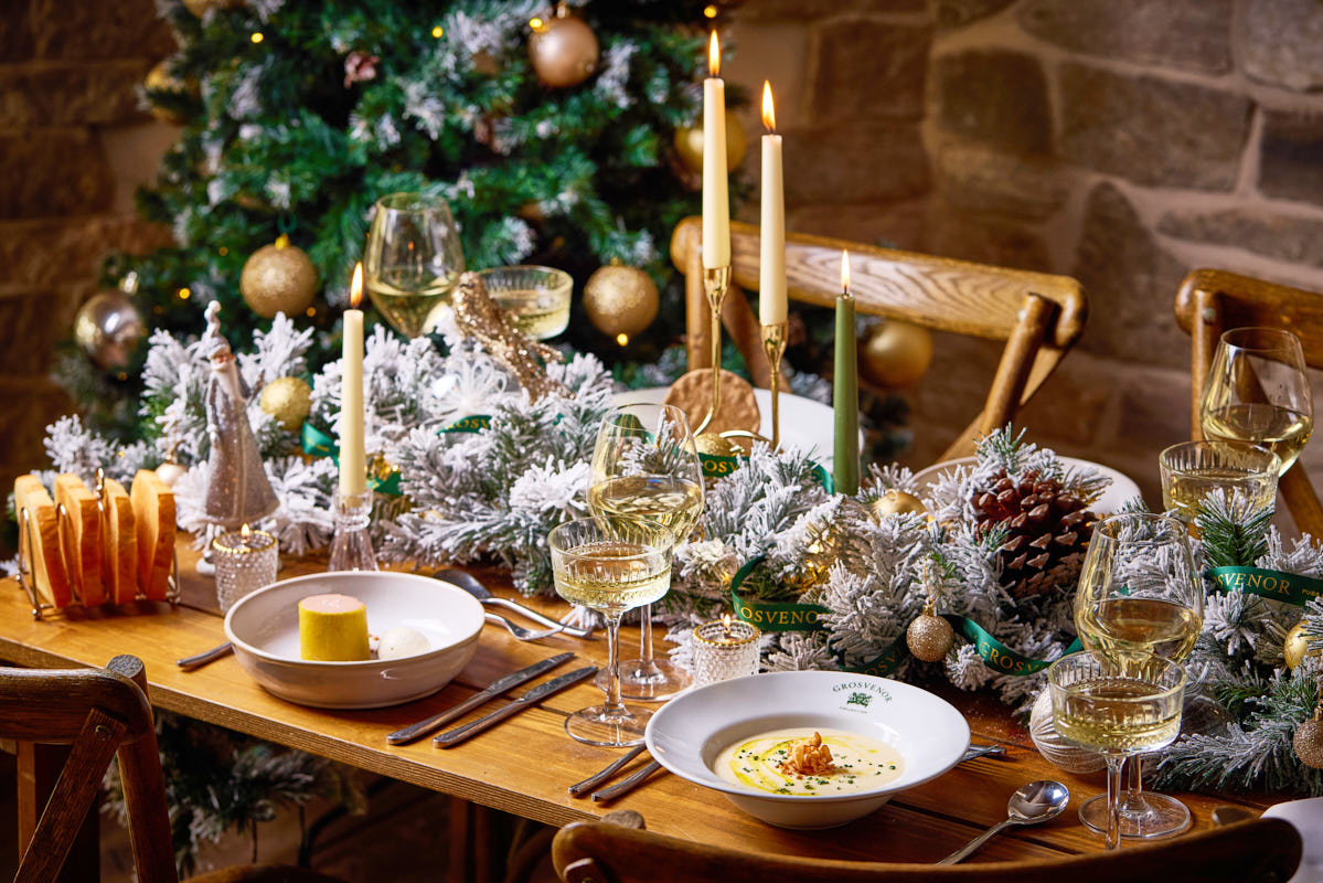 Festive dining table with elegant settings, candles, wine glasses, and Christmas decor, set against a lit Christmas tree backdrop. - Wild Thyme & Honey Hotel in the Cotswolds
