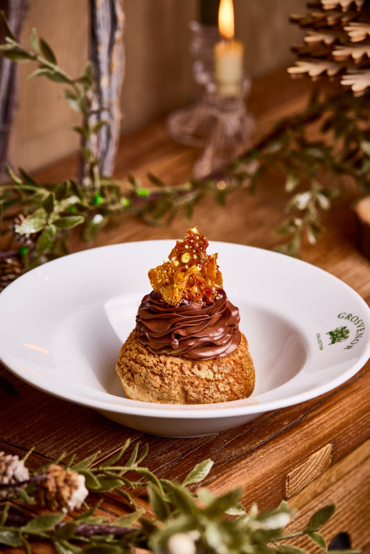 Cream puff topped with chocolate icing and caramel shards on a white plate, surrounded by rustic decor and greenery. - Wild Thyme & Honey Hotel in the Cotswolds