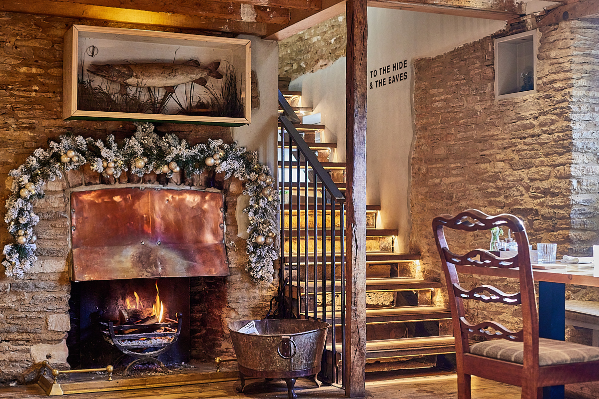 Rustic room with a lit fireplace, copper hood, and festive garland. Wooden staircase, stone walls, and cozy chairs complete the scene. - Wild Thyme & Honey Hotel in the Cotswolds