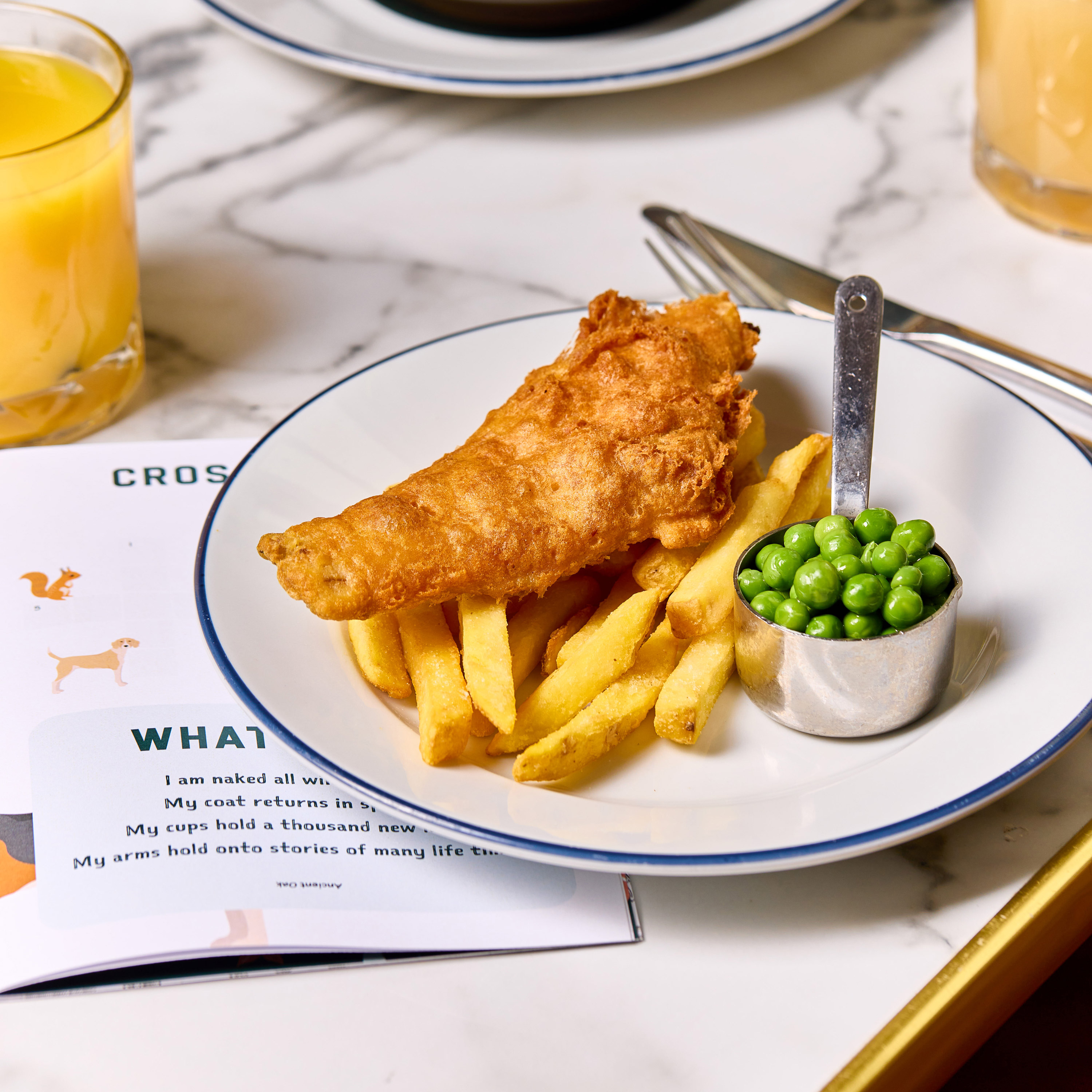 A plate of fish and chips with a side of peas on a marble table, accompanied by a crossword puzzle and orange juice. - Wild Thyme & Honey Hotel in the Cotswolds