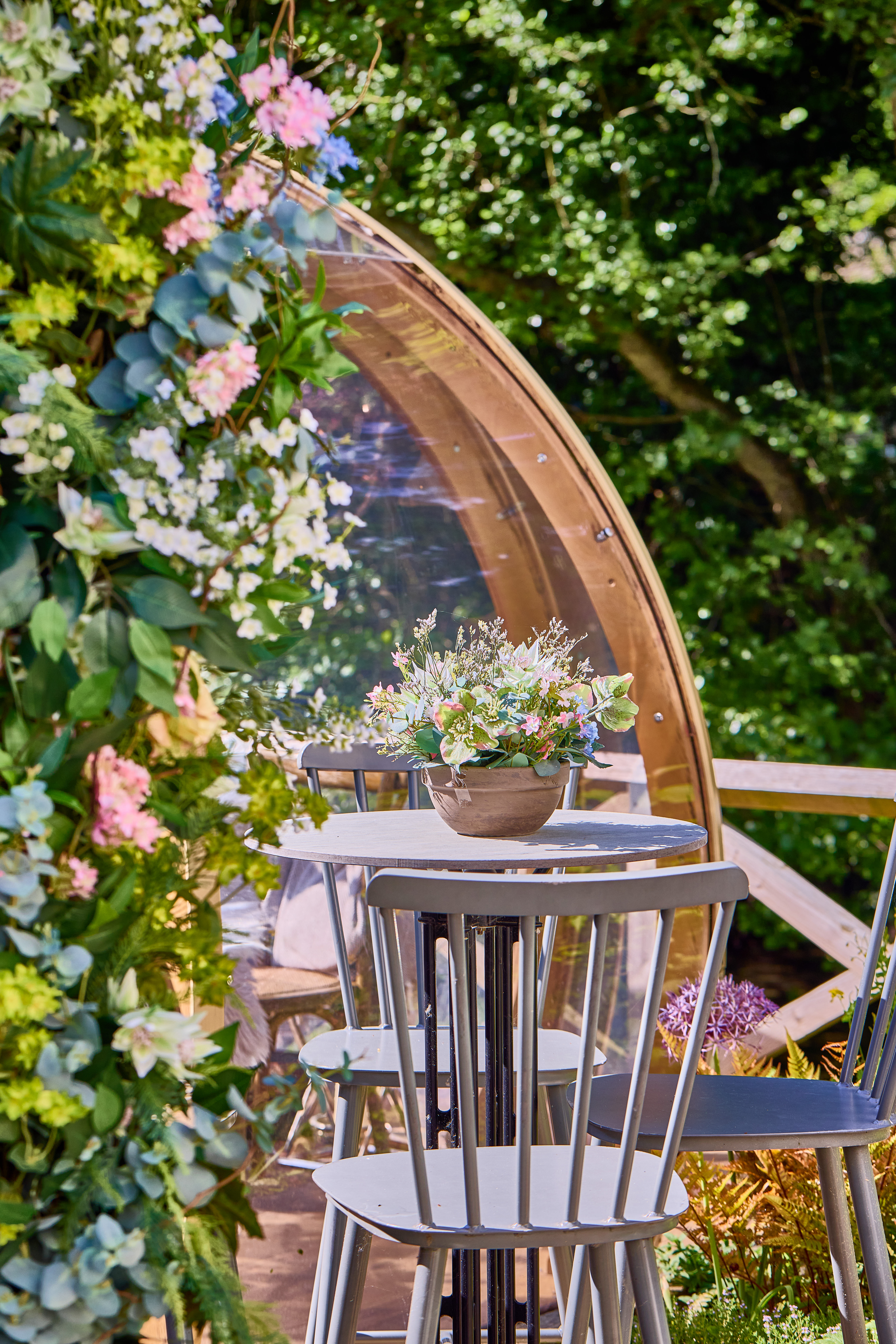 Outdoor dining area by a transparent dome, with a round table, chairs, and a flower arrangement, surrounded by greenery. - Wild Thyme & Honey Hotel in the Cotswolds