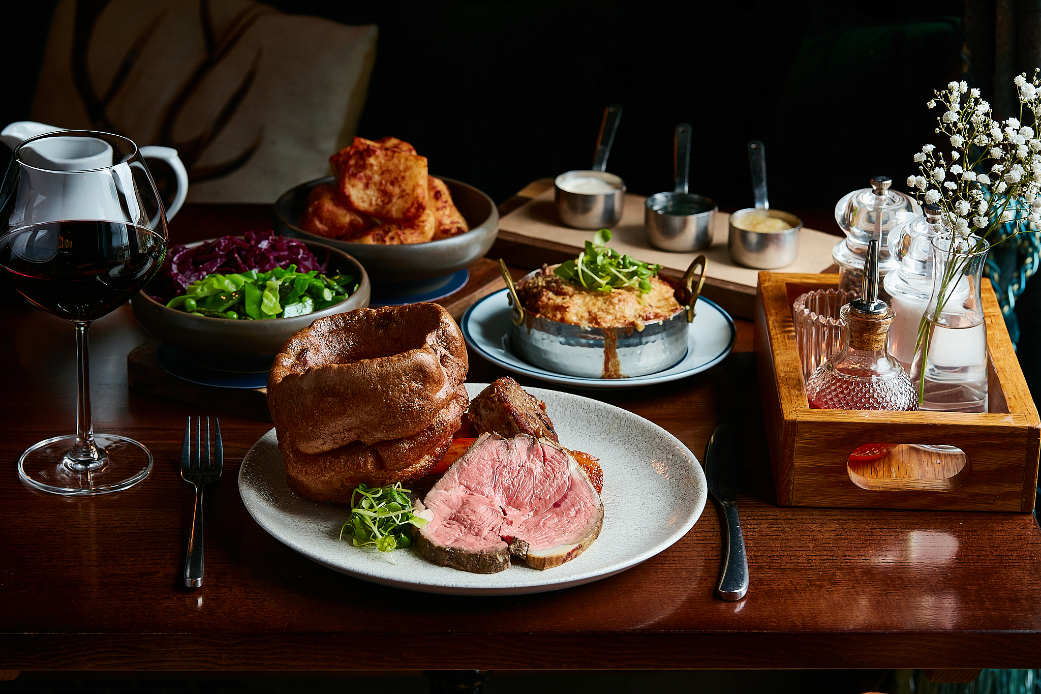 A plate with roast beef, Yorkshire pudding, and garnish, accompanied by wine, sides, condiments, and sauces on a wooden table. - Wild Thyme & Honey Hotel in the Cotswolds