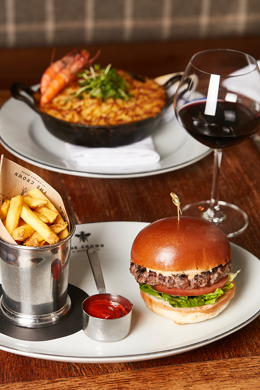 A gourmet meal featuring a juicy burger with lettuce on a bun, a side of fries in a metal cup with ketchup, a dish of seafood risotto topped with shrimp, and a glass of red wine on a wooden table. - Wild Thyme & Honey Hotel in the Cotswolds