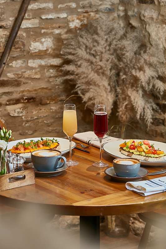 A wooden table set with two pizzas, two flutes of drinks, two cappuccinos, and a rustic brick wall in the background. - Wild Thyme & Honey Hotel in the Cotswolds