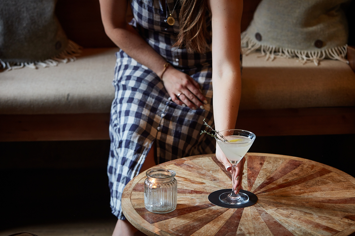 A person wearing a checkered dress reaches to place a cocktail on a circular wooden table. The table also holds a small lit candle in a glass holder. The background features a cushioned bench with pillows. - Wild Thyme & Honey Hotel in the Cotswolds