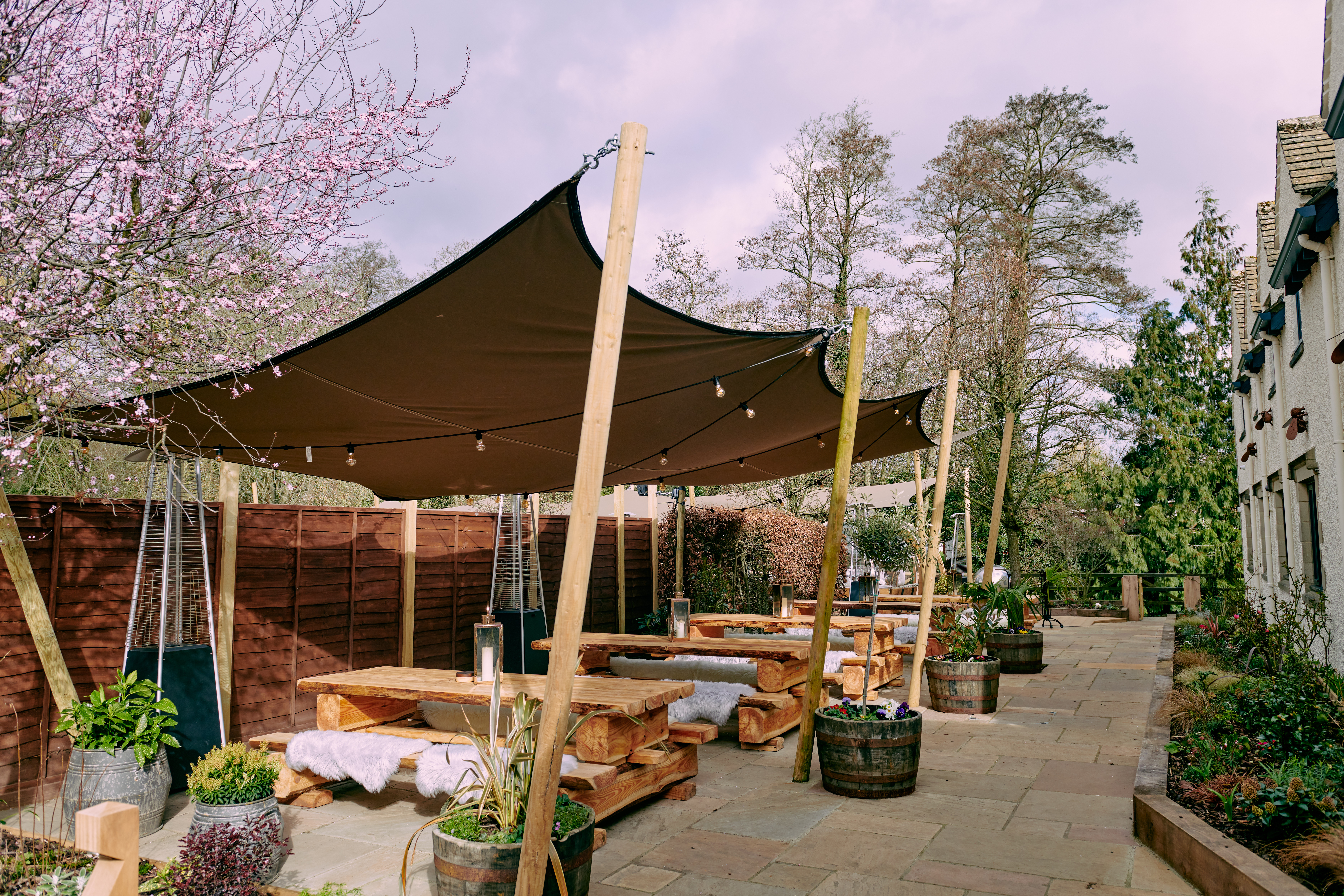 Outdoor seating area with wooden tables and benches under a large canopy, surrounded by trees and planters on a stone patio. - Wild Thyme & Honey Hotel in the Cotswolds