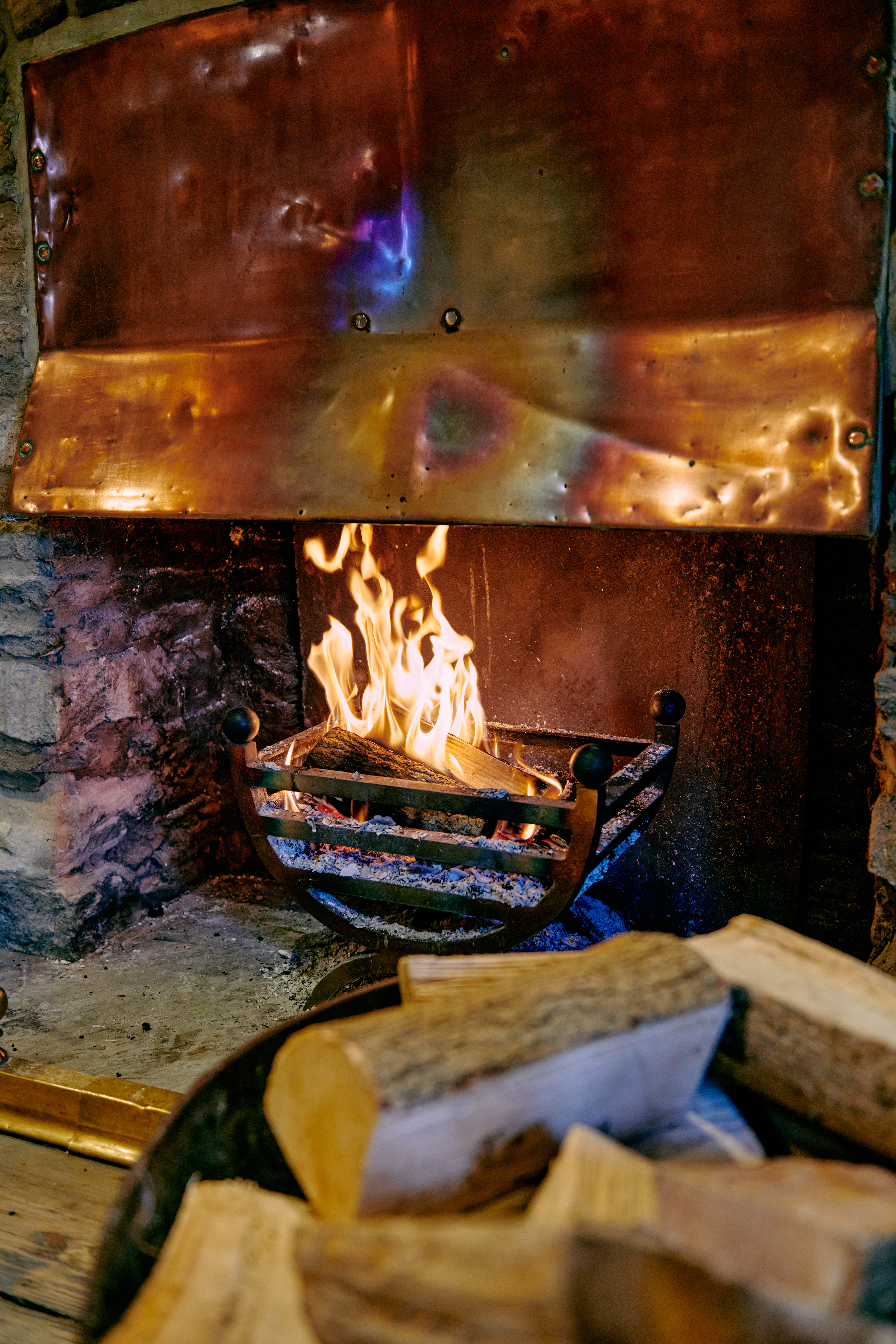 Lit fireplace with burning logs in a rustic setting, surrounded by a stone and copper design; extra logs in the foreground. - Wild Thyme & Honey Hotel in the Cotswolds