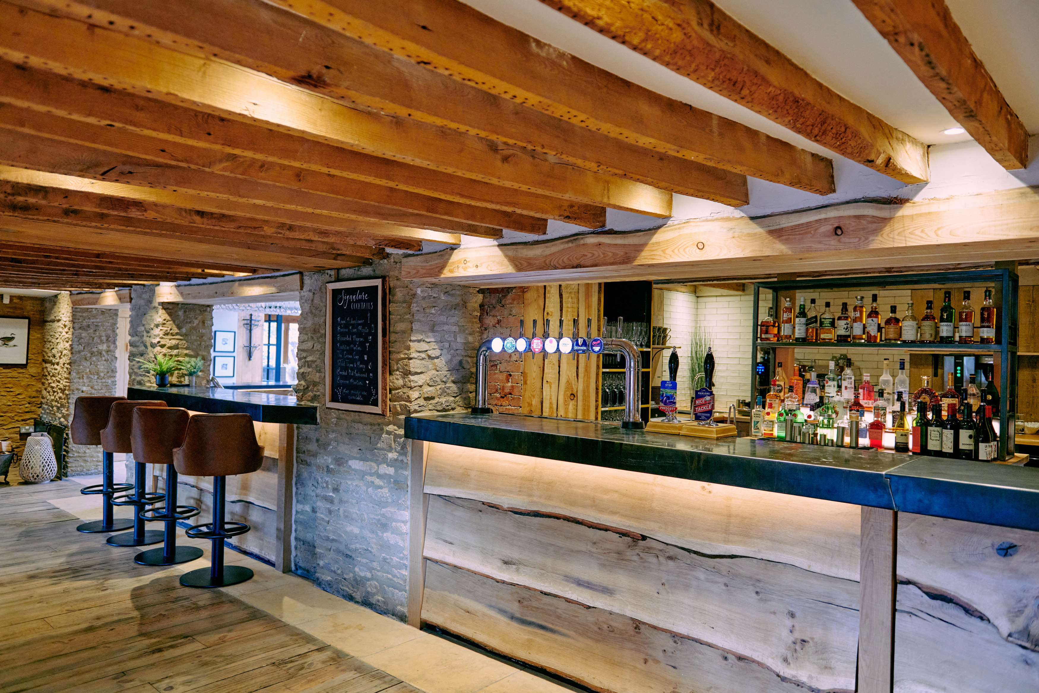 Rustic bar interior with exposed wooden beams and stone walls. Wooden bar counter with leather stools and shelves stocked with bottles. Soft lighting creates a cozy ambiance. A chalkboard menu hangs on the wall. - Wild Thyme & Honey Hotel in the Cotswolds