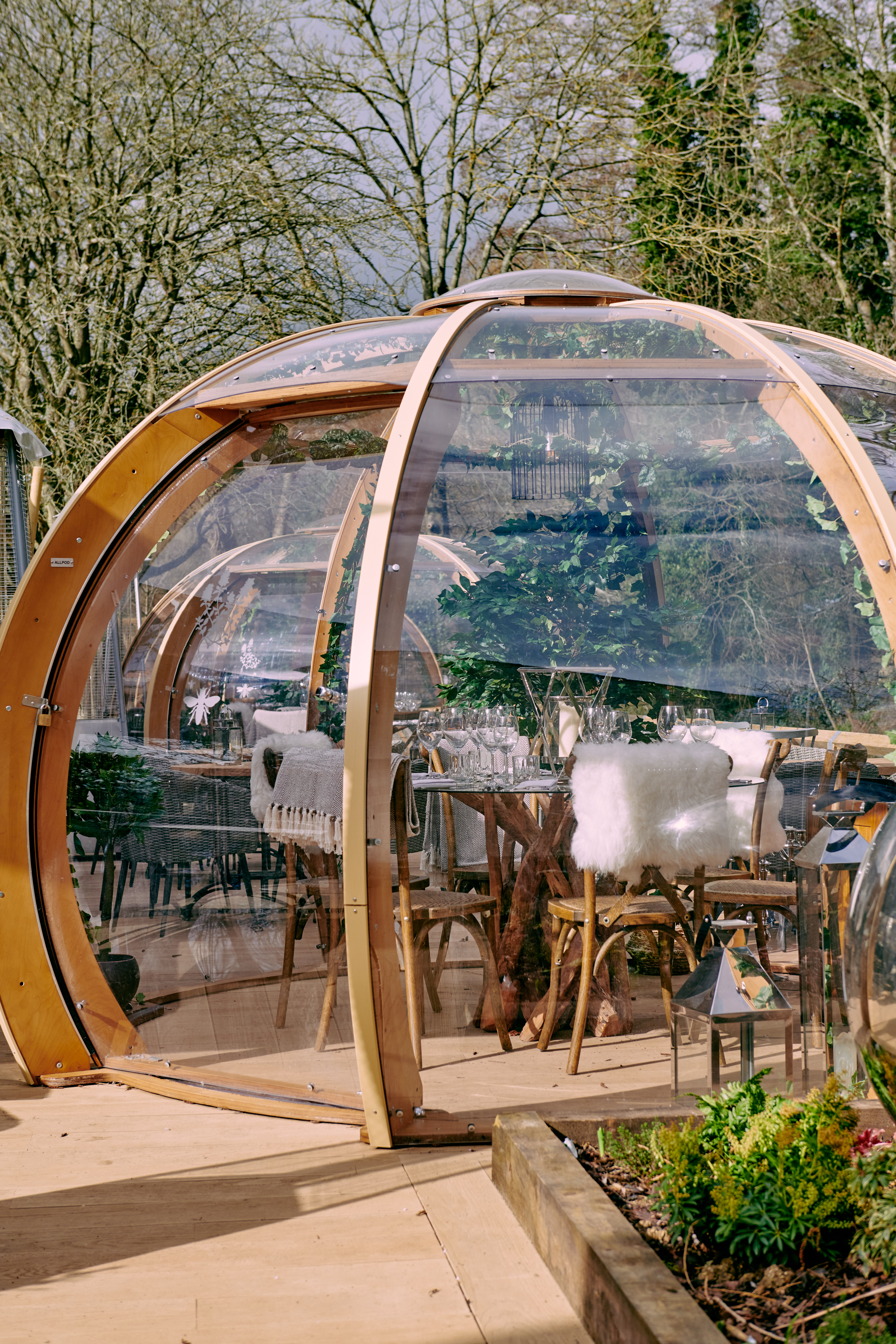 Glass igloo-style outdoor dining space with wooden arches and cozy decor, set amidst a garden with trees in the background. - Wild Thyme & Honey Hotel in the Cotswolds