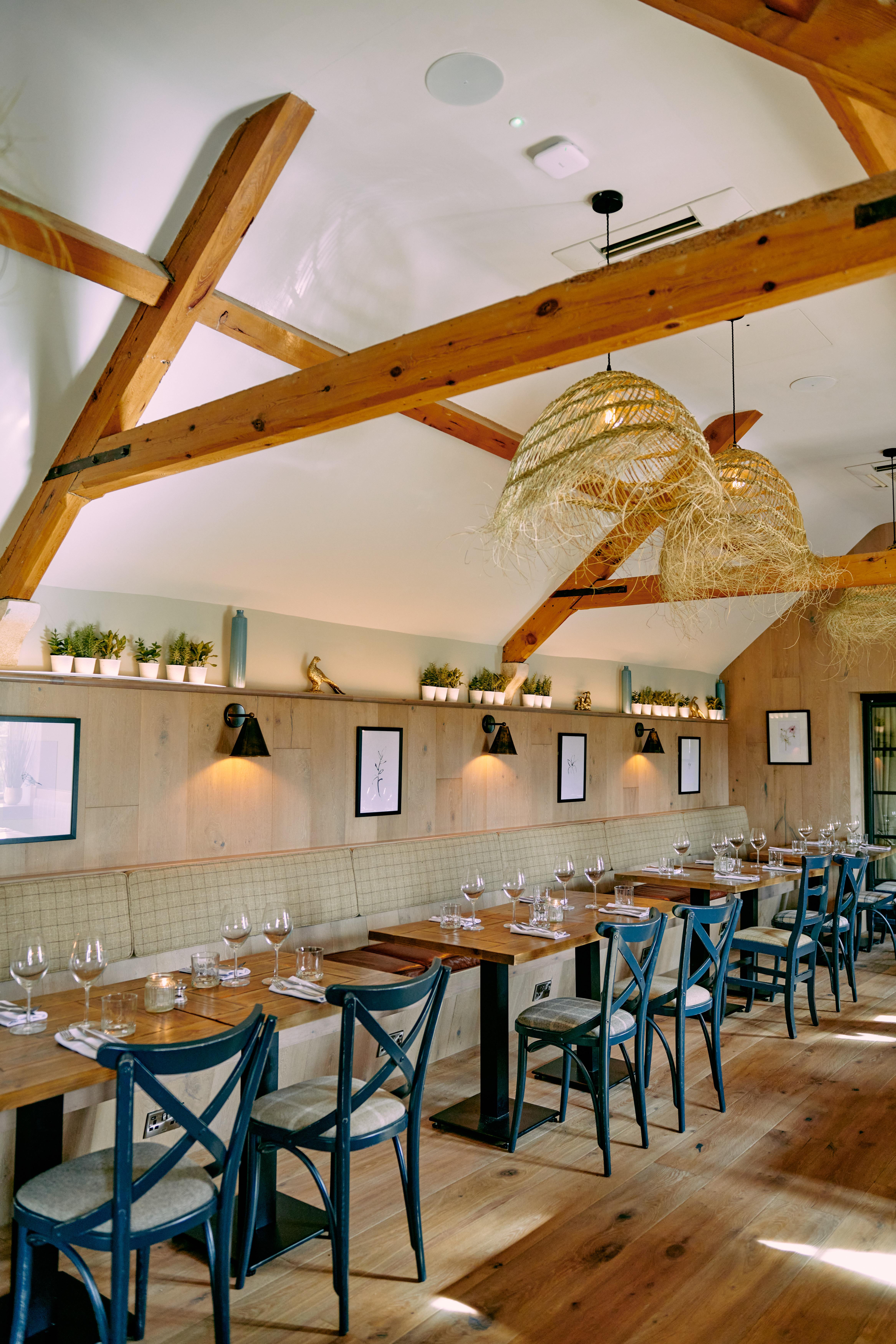 Rustic dining area with wooden beams, woven hanging lights, wooden tables and chairs, and wall shelving with plants. - Wild Thyme & Honey Hotel in the Cotswolds