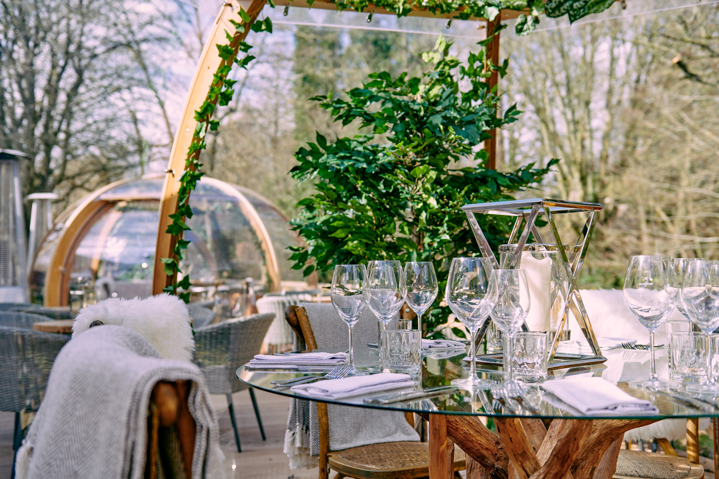 Outdoor dining area with a rustic glass table set elegantly, surrounded by greenery and a transparent dome structure in the background. - Wild Thyme & Honey Hotel in the Cotswolds