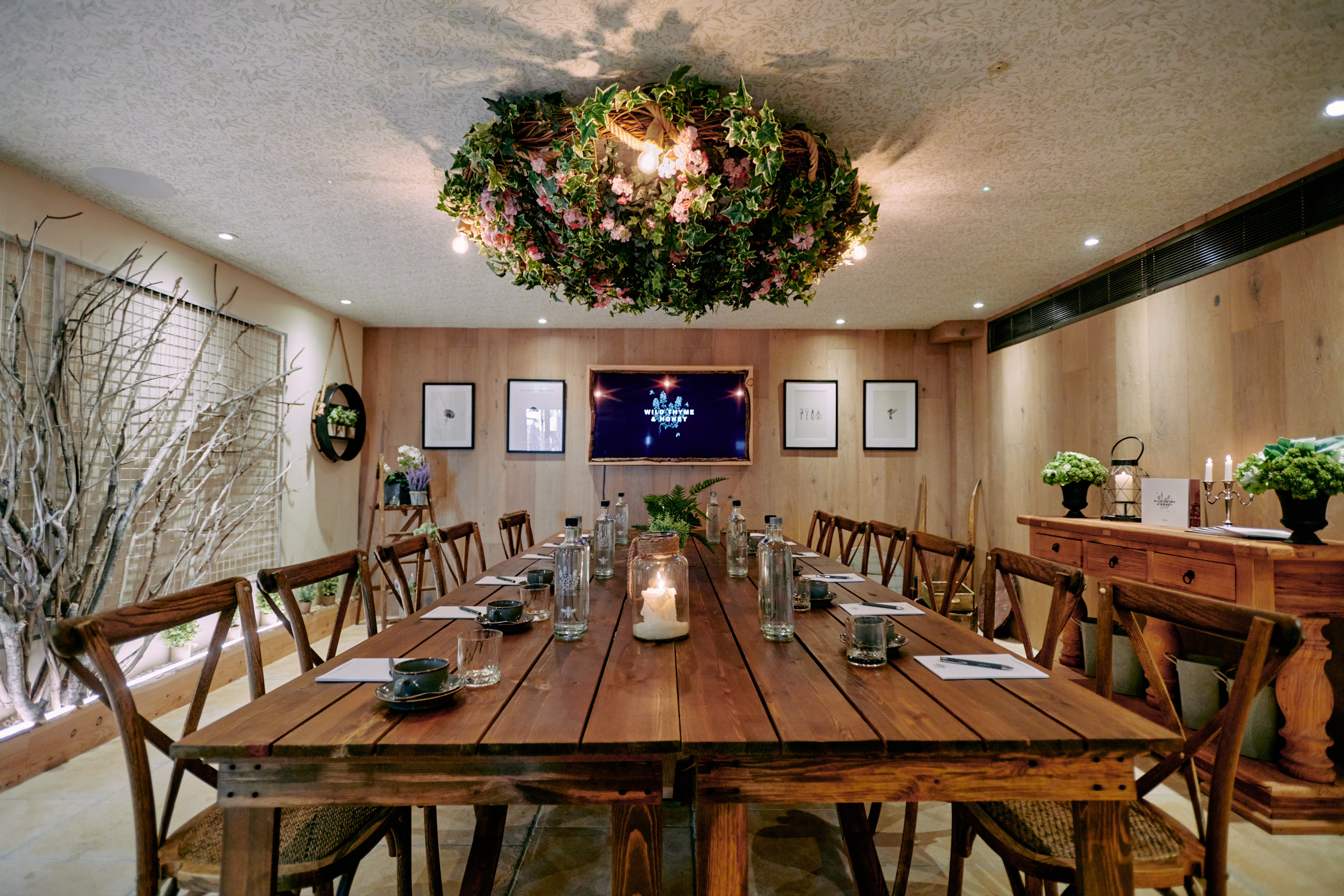 A rustic meeting room with a wooden table, chairs, a floral chandelier, and framed art on wood-paneled walls. - Wild Thyme & Honey Hotel in the Cotswolds