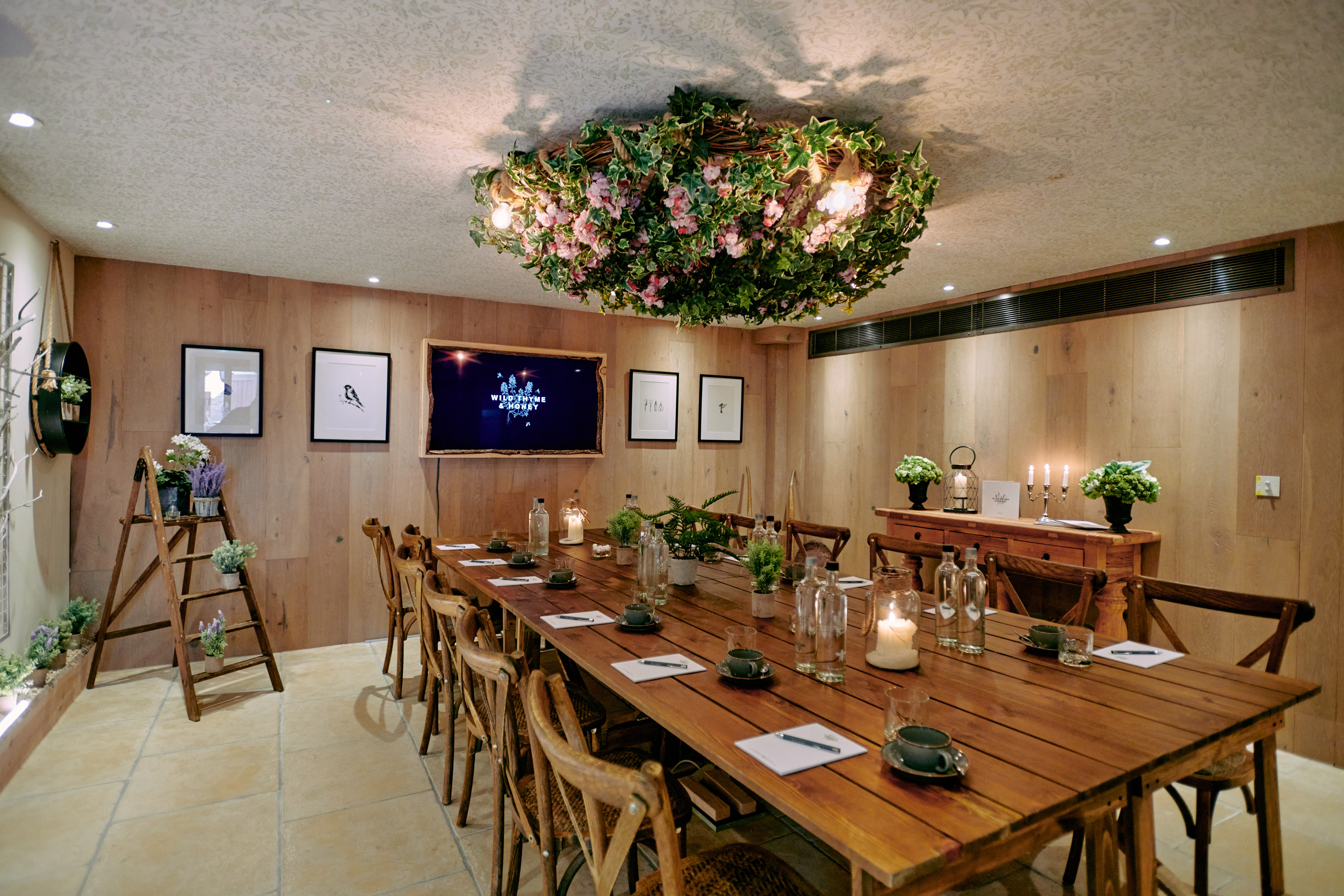 A cozy dining room with a long wooden table, decorated with candles and plants. A floral chandelier hangs above. - Wild Thyme & Honey Hotel in the Cotswolds