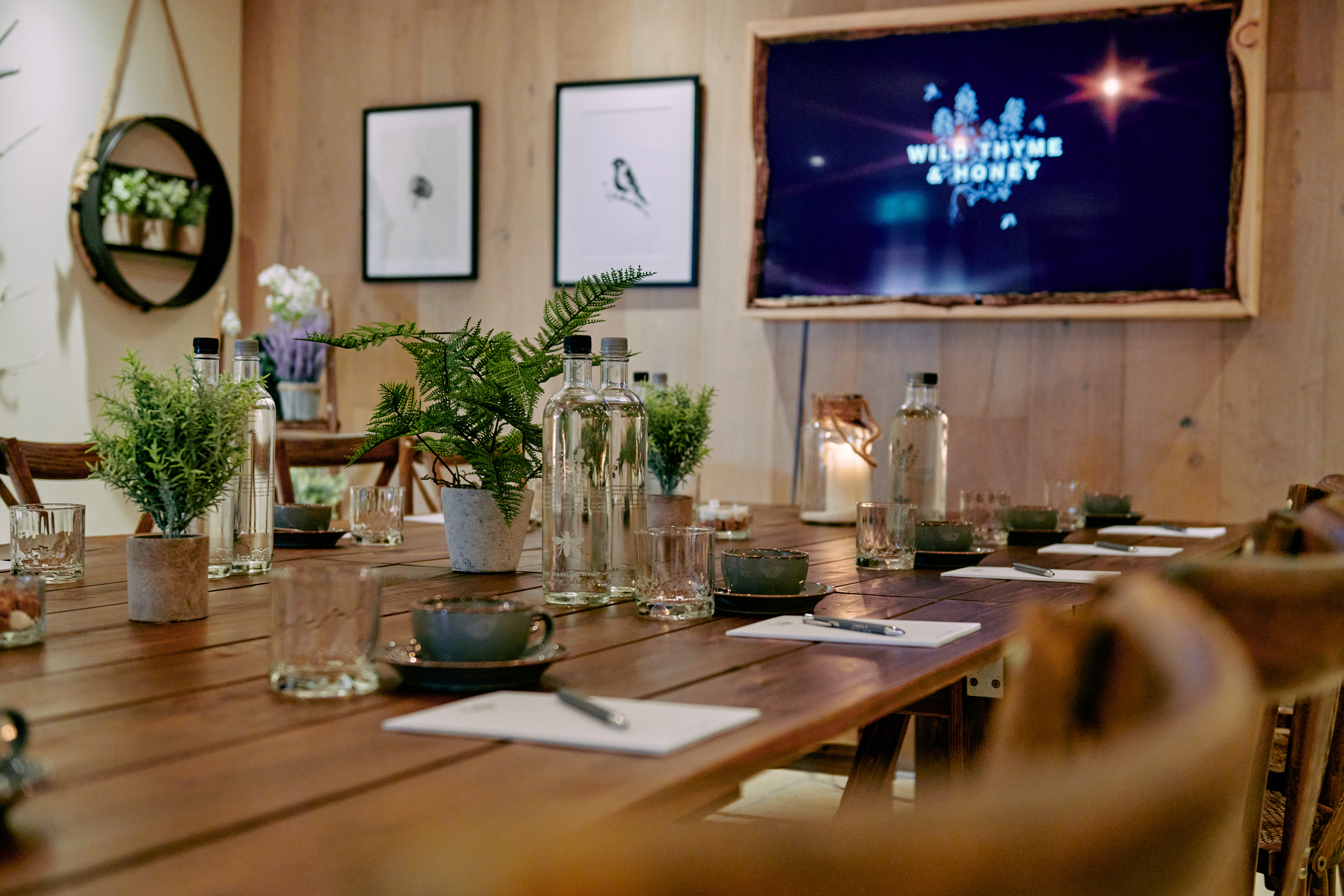 Conference room with a wooden table, notebooks, bottles, plants, and a screen displaying Wild Thyme & Honey. - Wild Thyme & Honey Hotel in the Cotswolds
