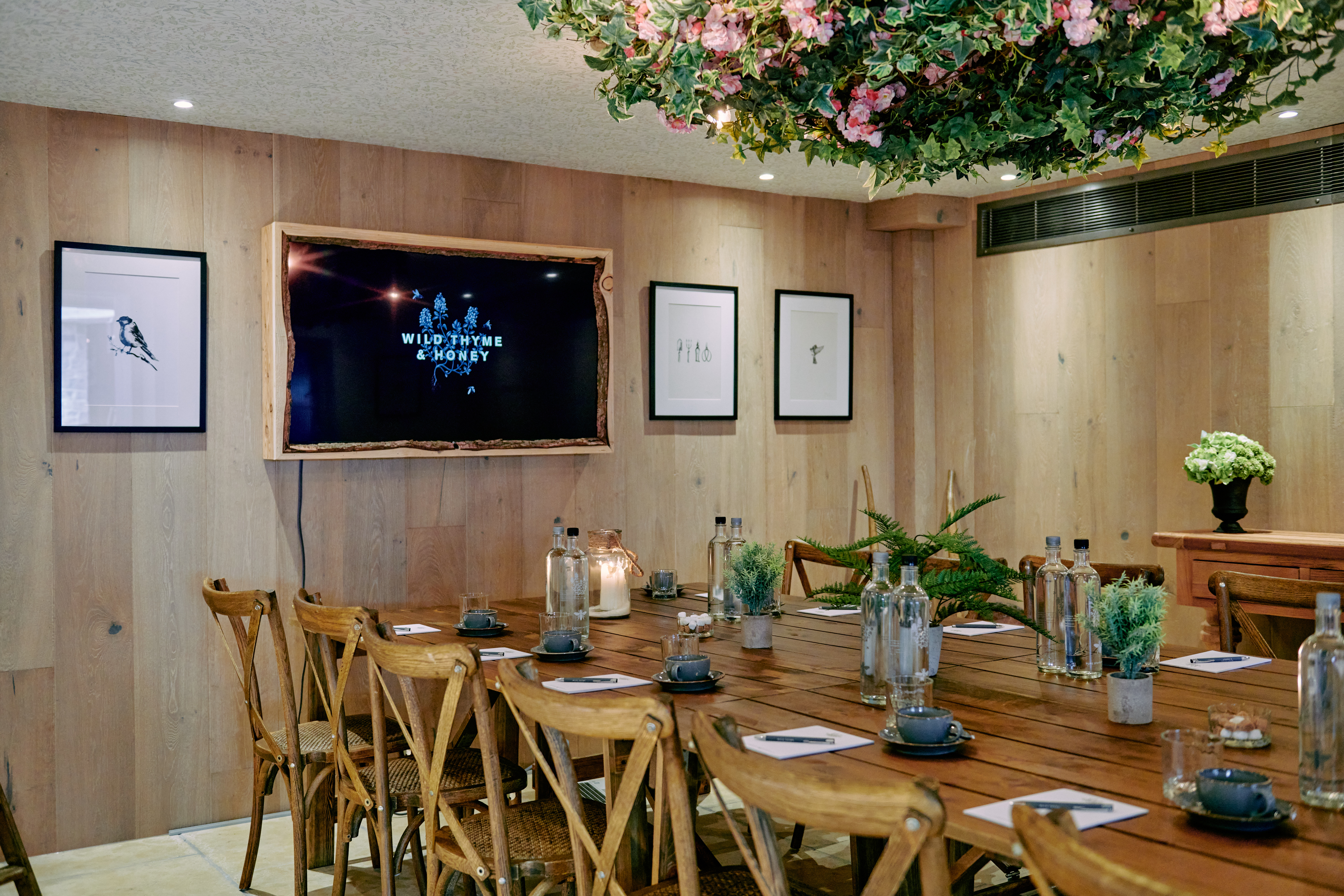 Cozy meeting room with wooden table, chairs, TV, framed art, and hanging floral decor. Water bottles and notebooks on table. - Wild Thyme & Honey Hotel in the Cotswolds