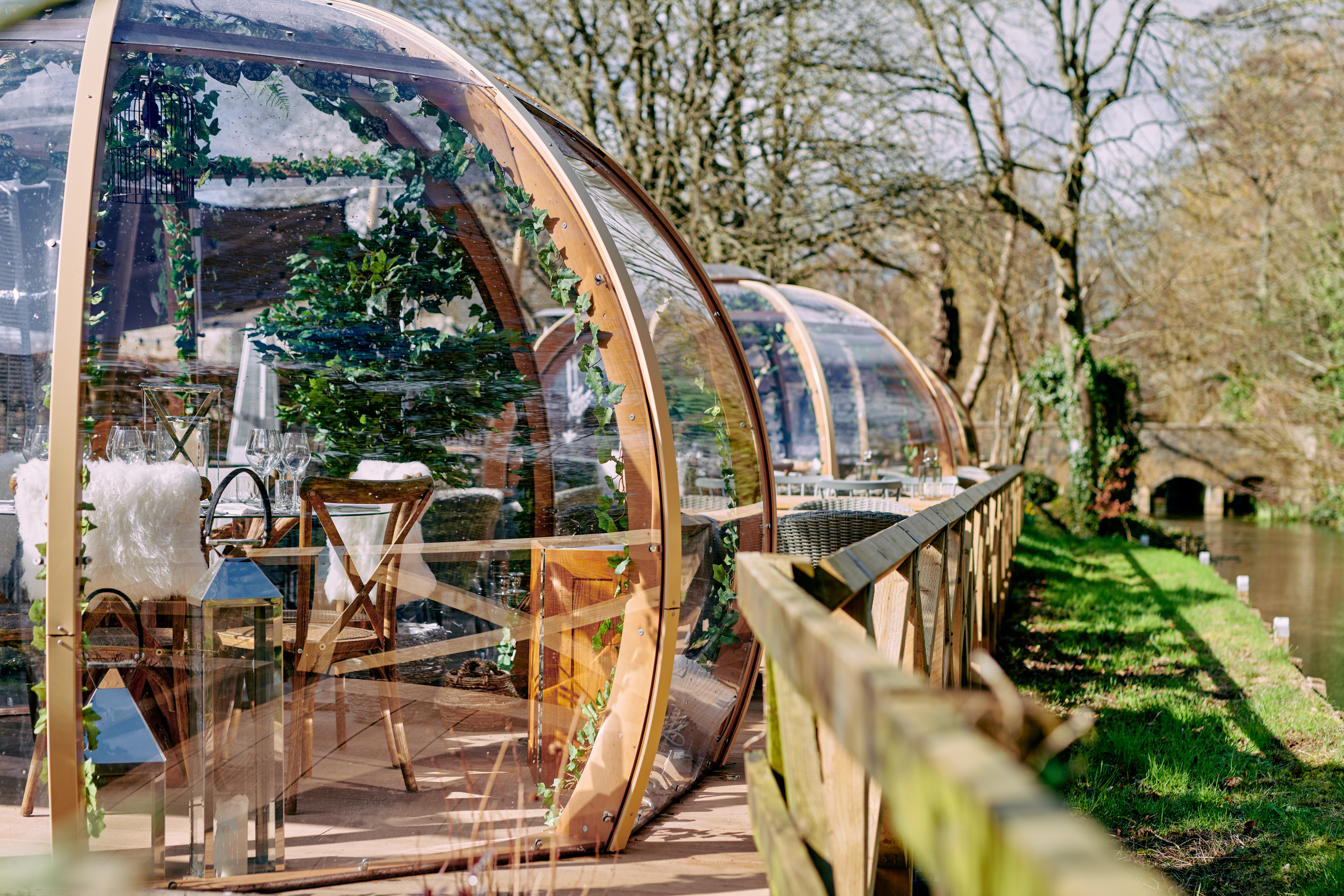 Glass domes along a riverbank with chairs and tables inside, surrounded by greenery and trees in the background. - Wild Thyme & Honey Hotel in the Cotswolds