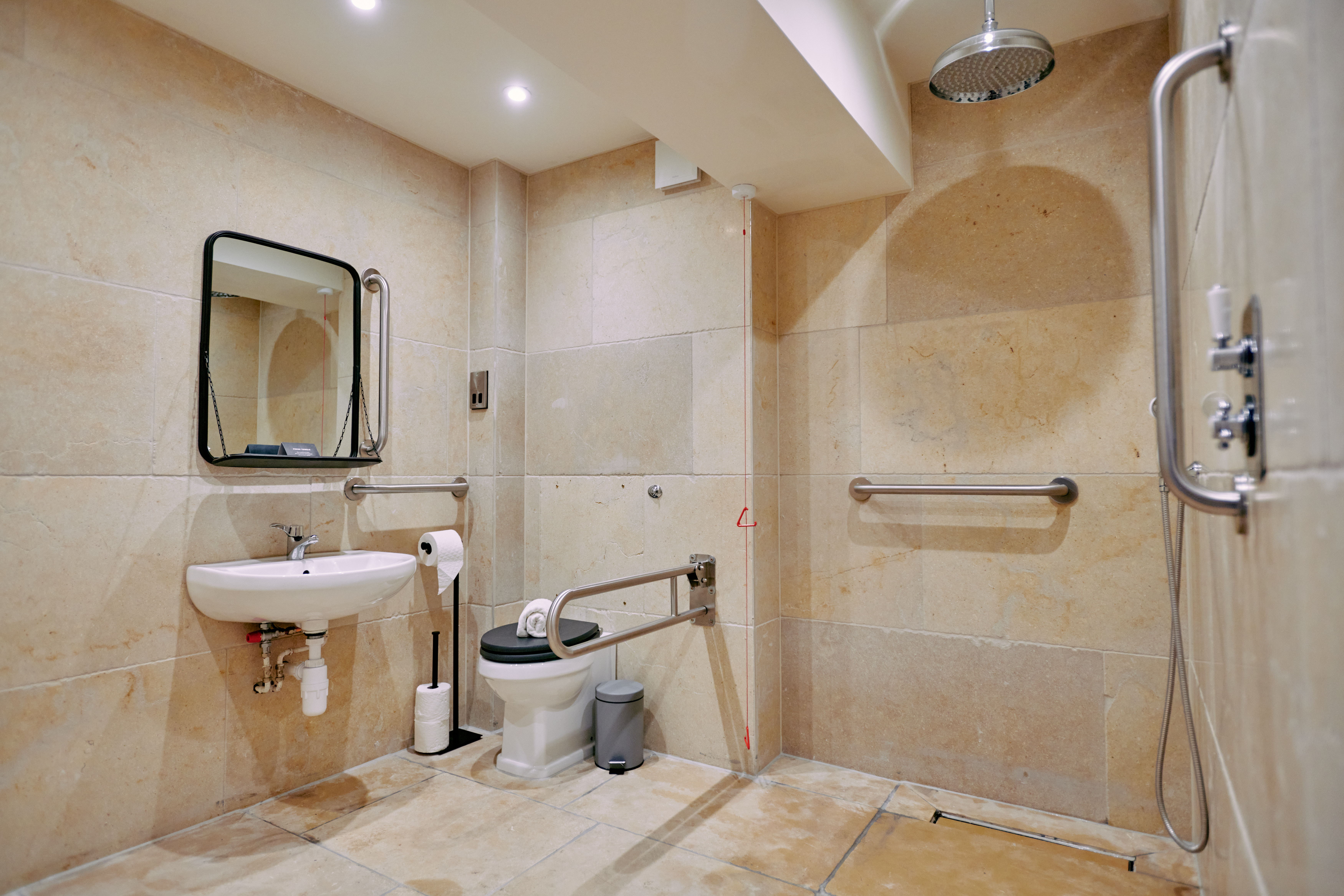 Accessible bathroom with beige tiles, equipped with a shower, toilet, grab bars, foldable support rail, and a small sink with a mirror. - Wild Thyme & Honey Hotel in the Cotswolds