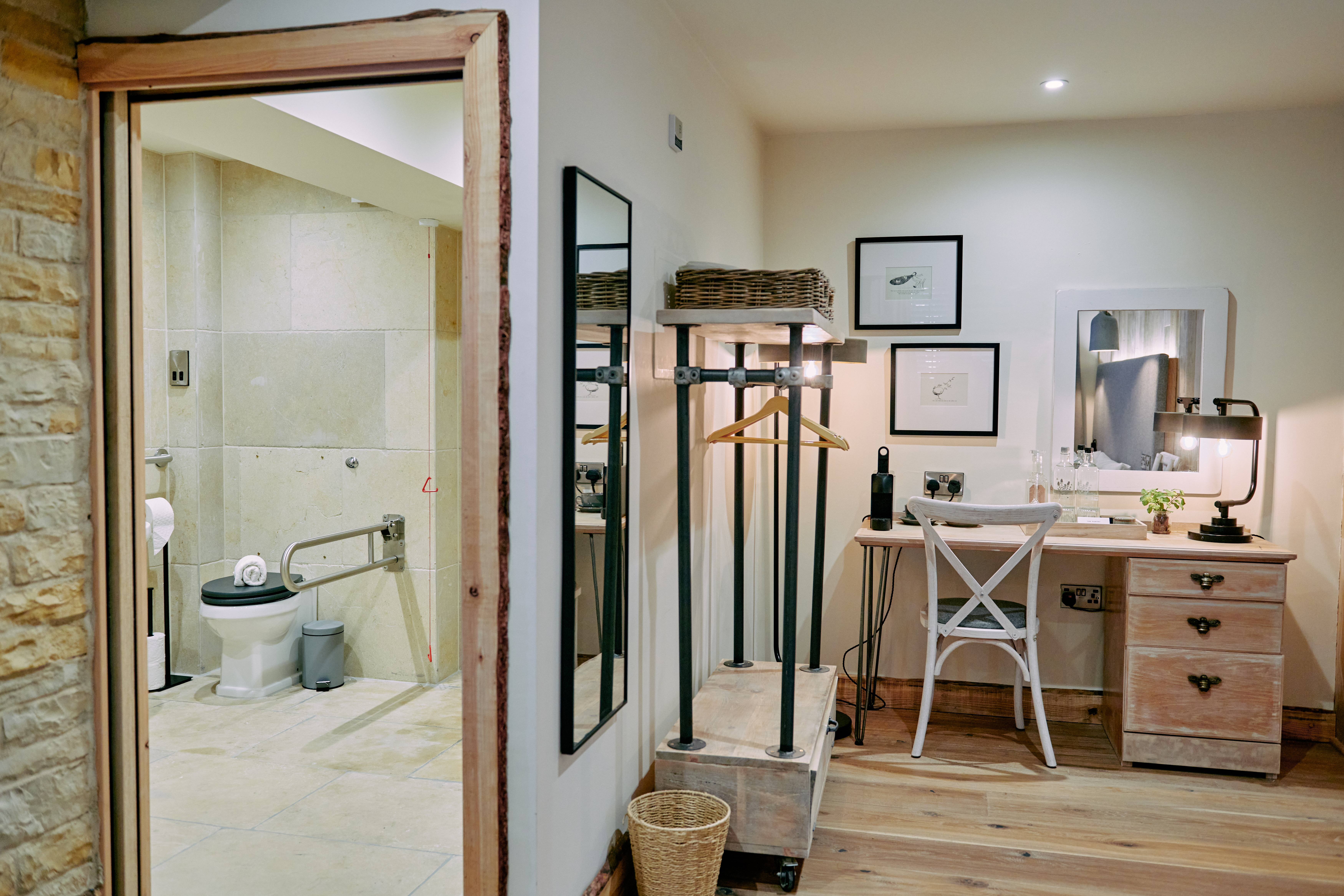 A dressing area with a vanity table and chair next to a bathroom with accessible features, like a grab bar near the toilet. - Wild Thyme & Honey Hotel in the Cotswolds