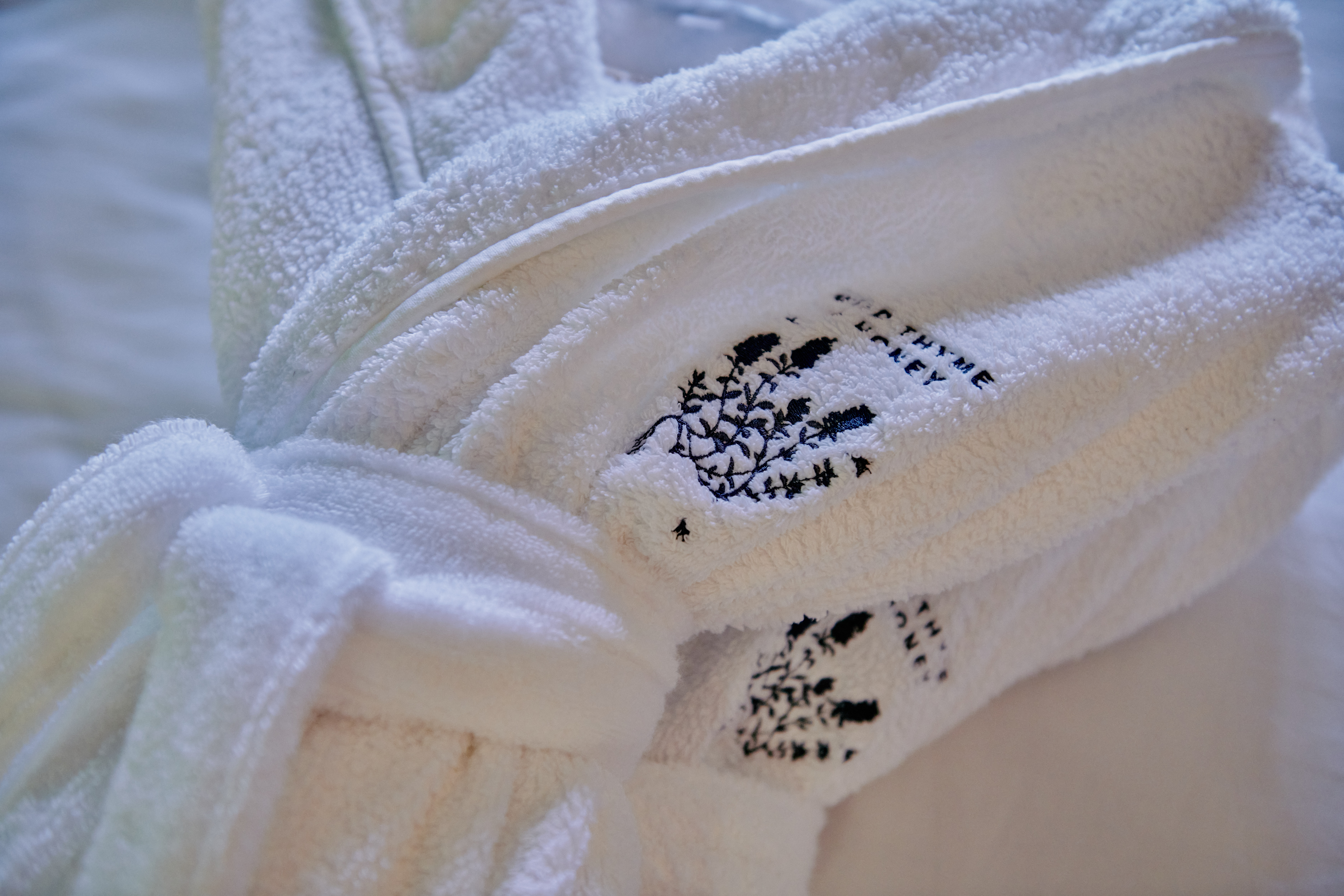 Close-up of a folded white bathrobe with embroidered black tree designs, resting on a soft, light-colored surface. - Wild Thyme & Honey Hotel in the Cotswolds