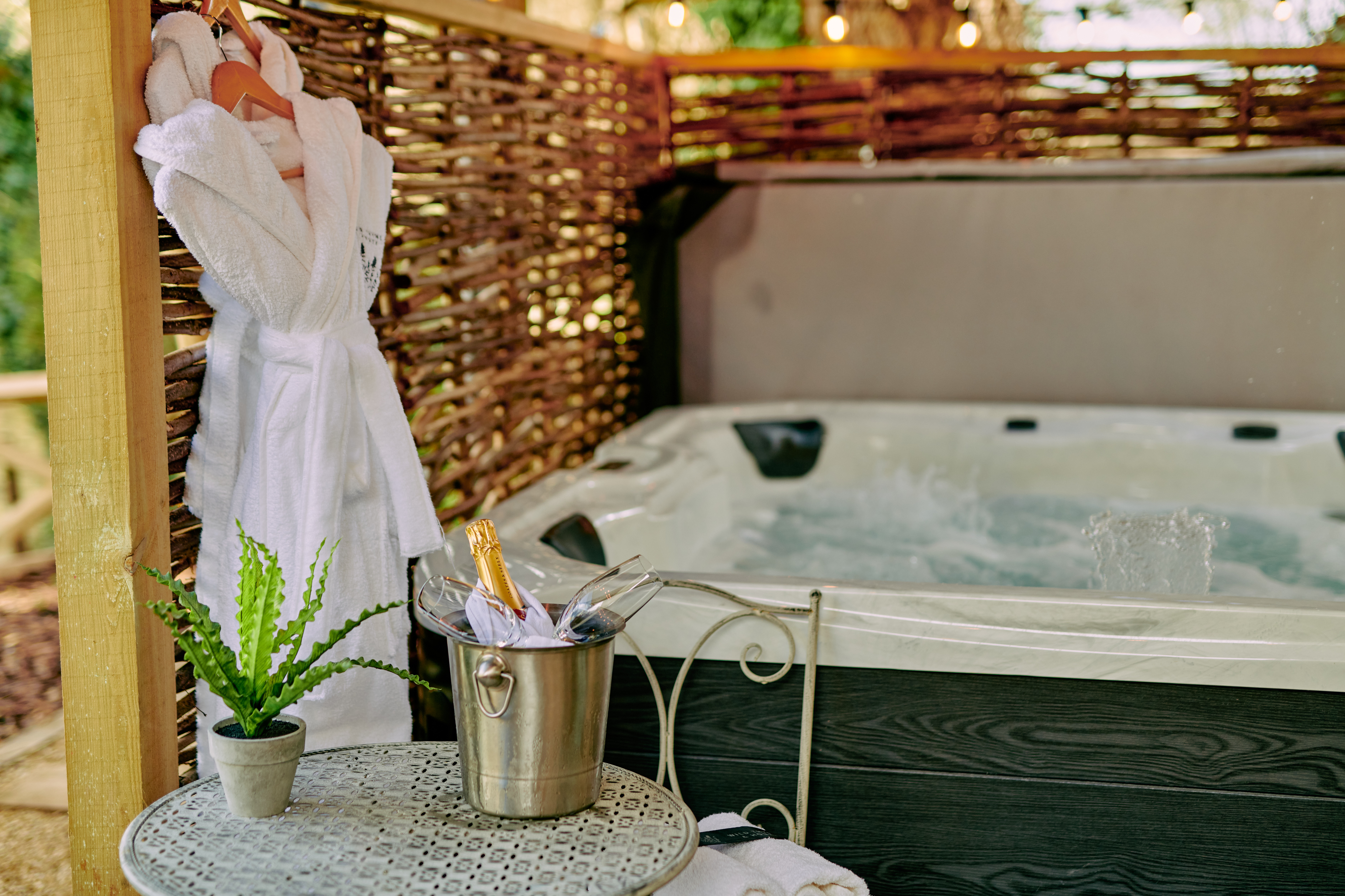 Private hot tub area with a frothy jacuzzi, a bucket of champagne on a table, a potted plant, and a hanging robe outside. - Wild Thyme & Honey Hotel in the Cotswolds