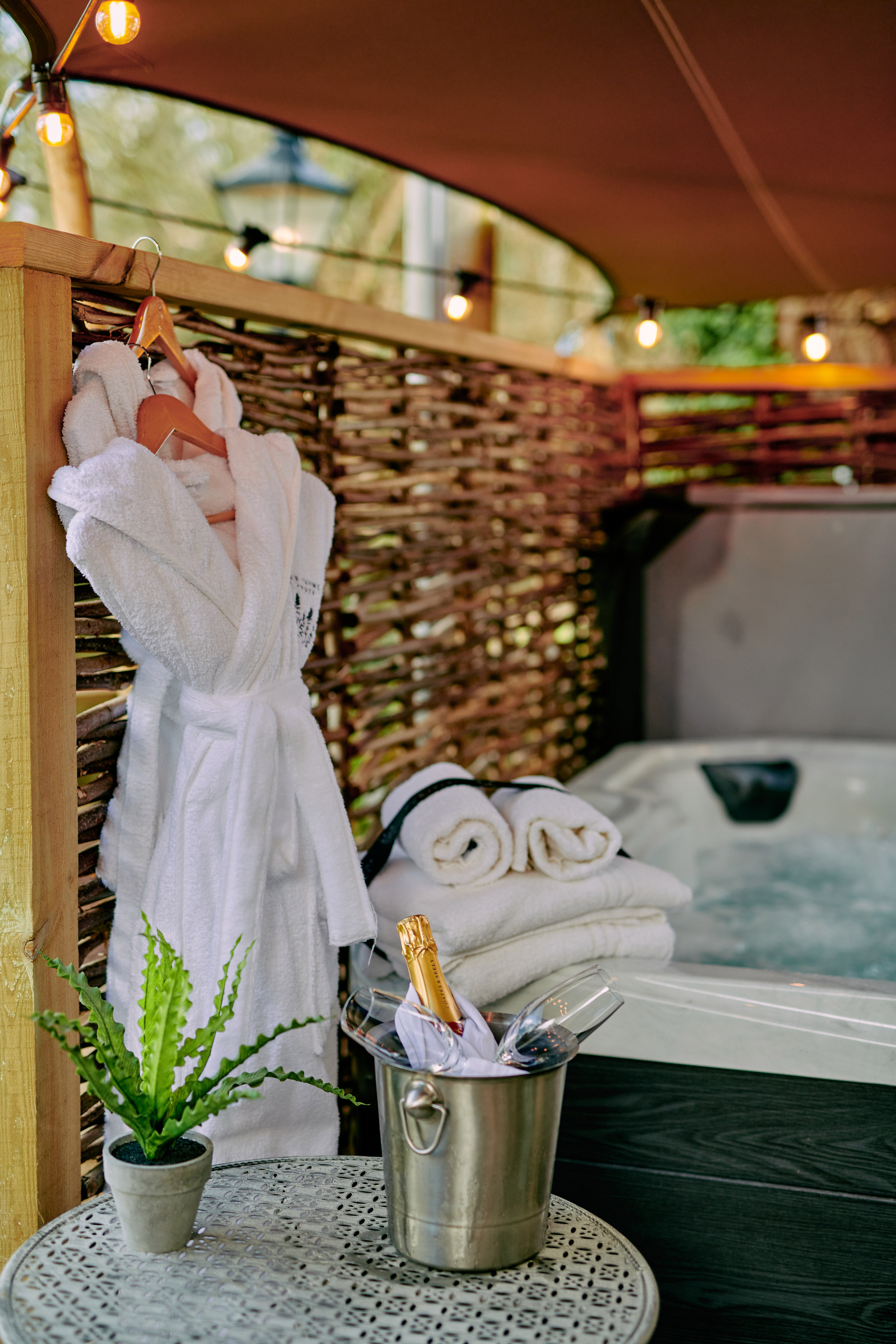 Outdoor spa setup with a hot tub, folded towels, and a hanging robe. A champagne bottle in an ice bucket sits on a small table. - Wild Thyme & Honey Hotel in the Cotswolds