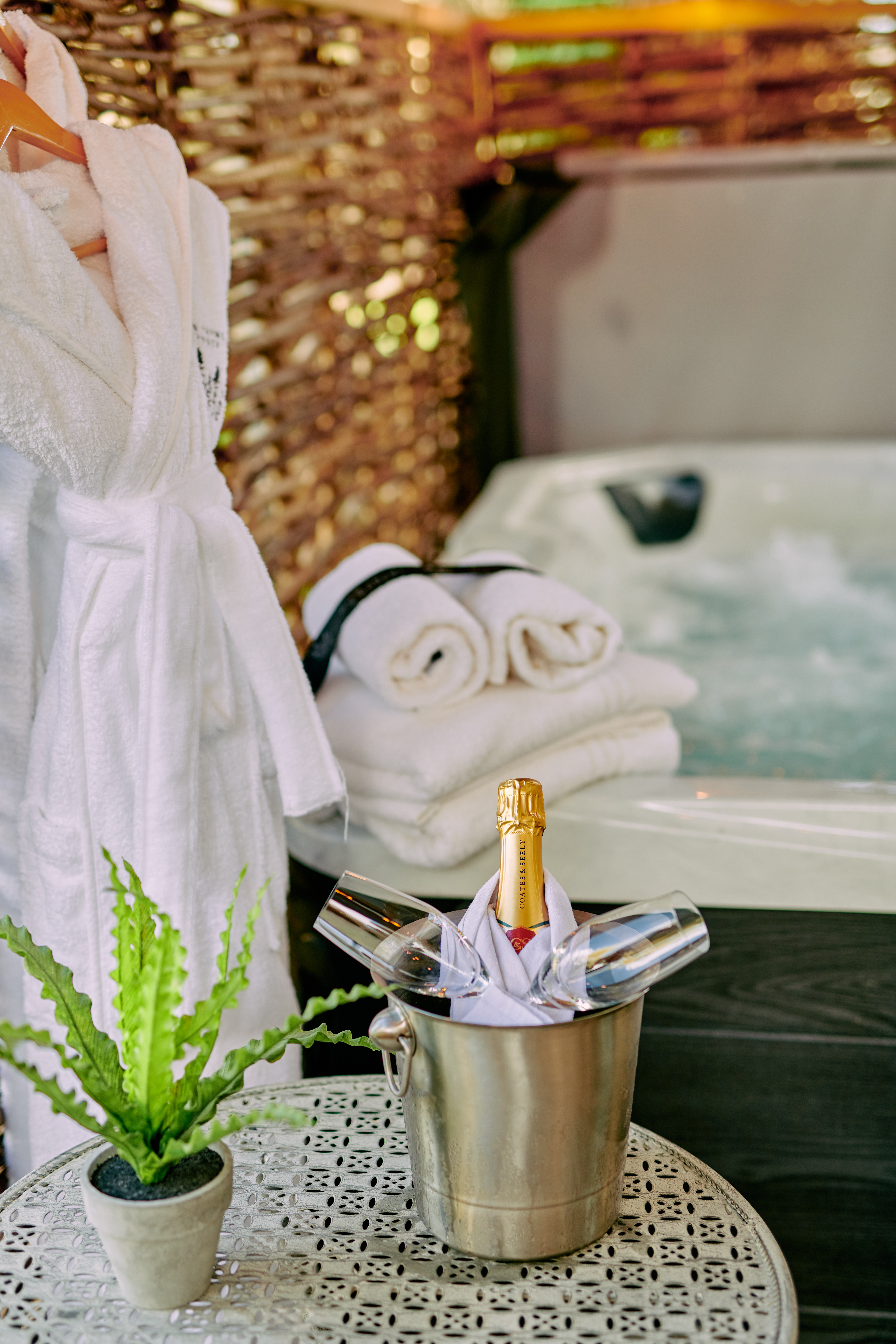 Champagne in an ice bucket and towels on a table beside a bubbling hot tub, with a small potted plant nearby. - Wild Thyme & Honey Hotel in the Cotswolds