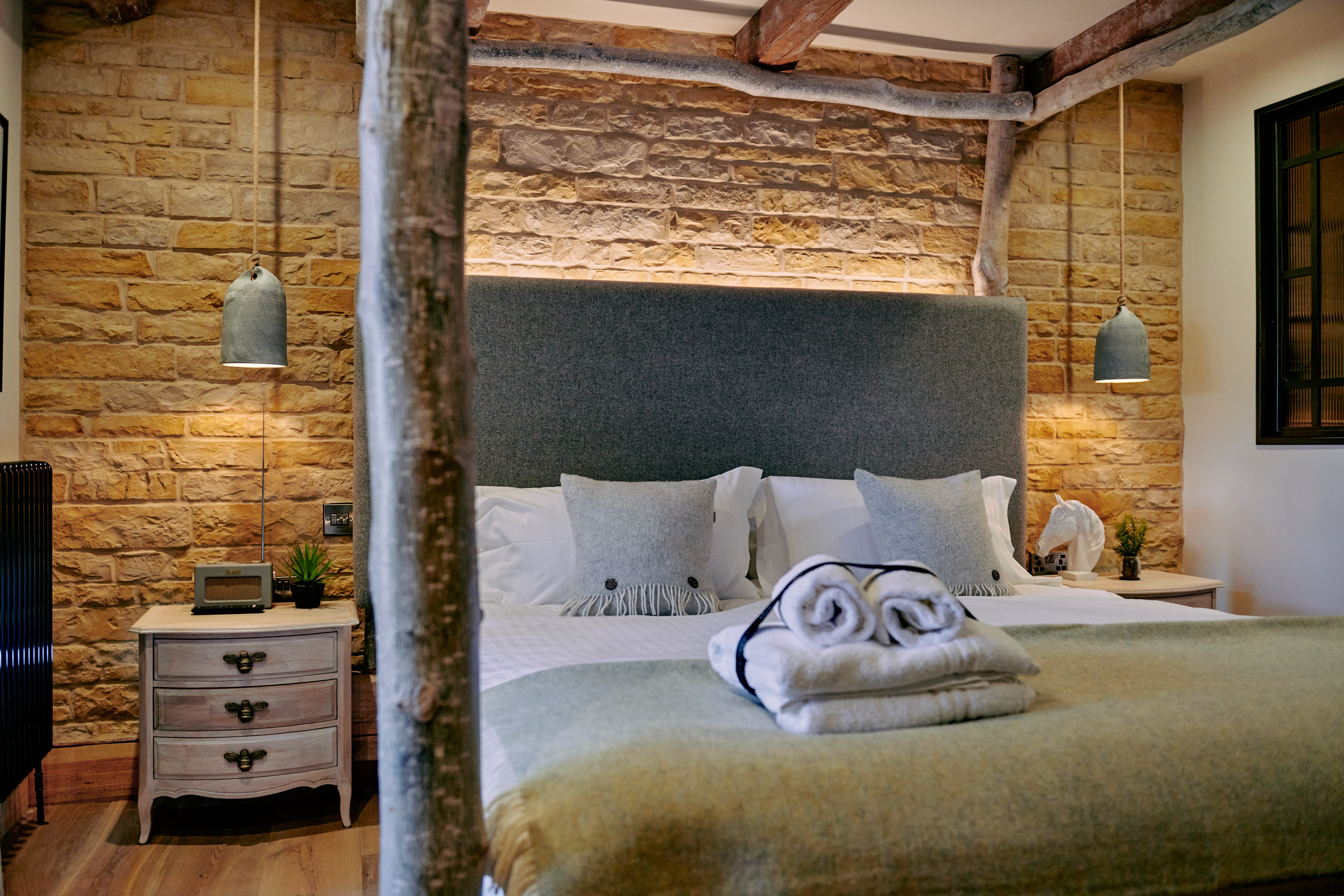 Cozy bedroom with a double bed, stone walls, wooden beams, a soft gray headboard, and folded towels on the foot of the bed. - Wild Thyme & Honey Hotel in the Cotswolds