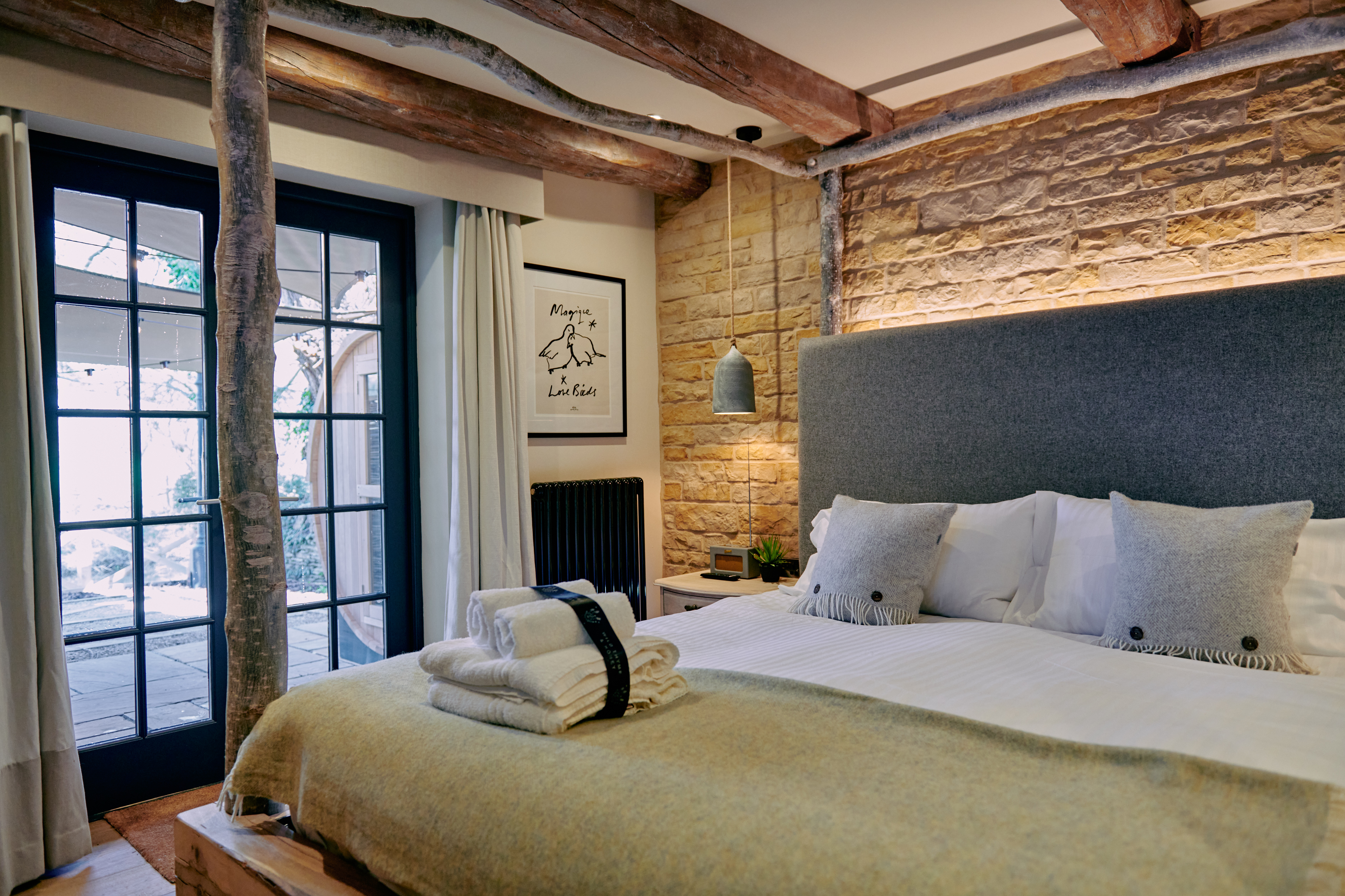 Cozy bedroom with a rustic design, features a bed with a gray headboard, stone walls, wooden beams, and a door to a patio. - Wild Thyme & Honey Hotel in the Cotswolds